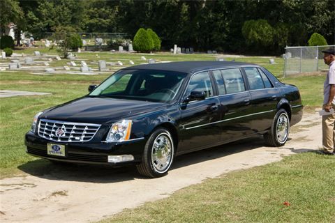 Black luxury sedan parked on a cemetery driveway beside gravestones and a man in tan pants