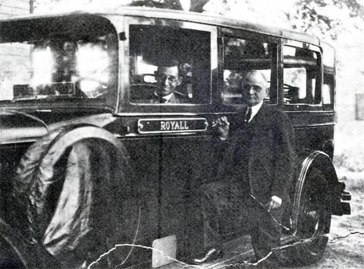 Man in a suit standing by a vintage Royal bus, with passengers visible inside.
