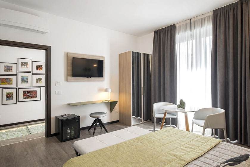 Habitación de hotel con cama, escritorio, sillas, TV y puerta abierta al pasillo con obras de arte enmarcadas.