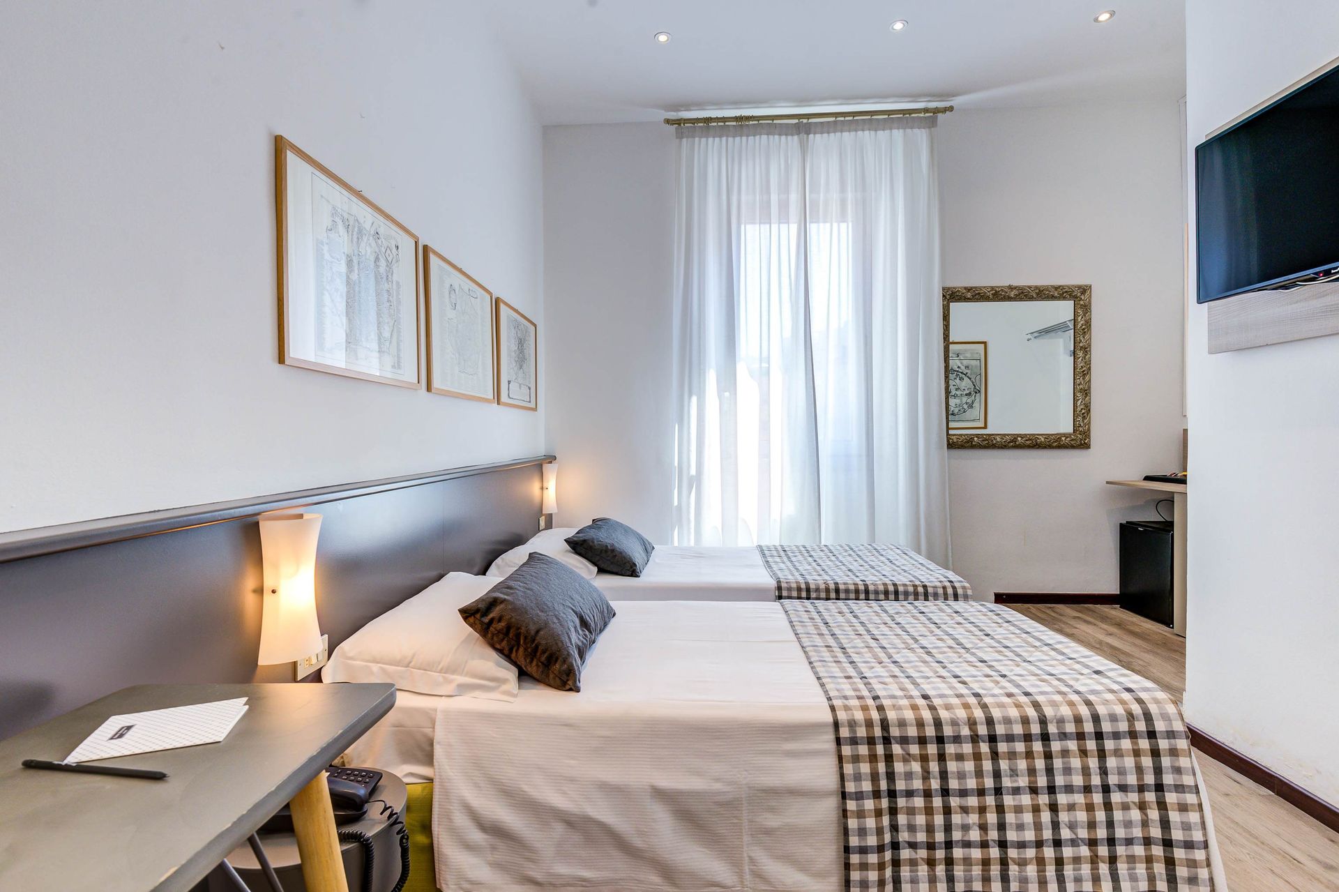 Dos camas individuales en una luminosa habitación de hotel con ventana, espejo y obras de arte.