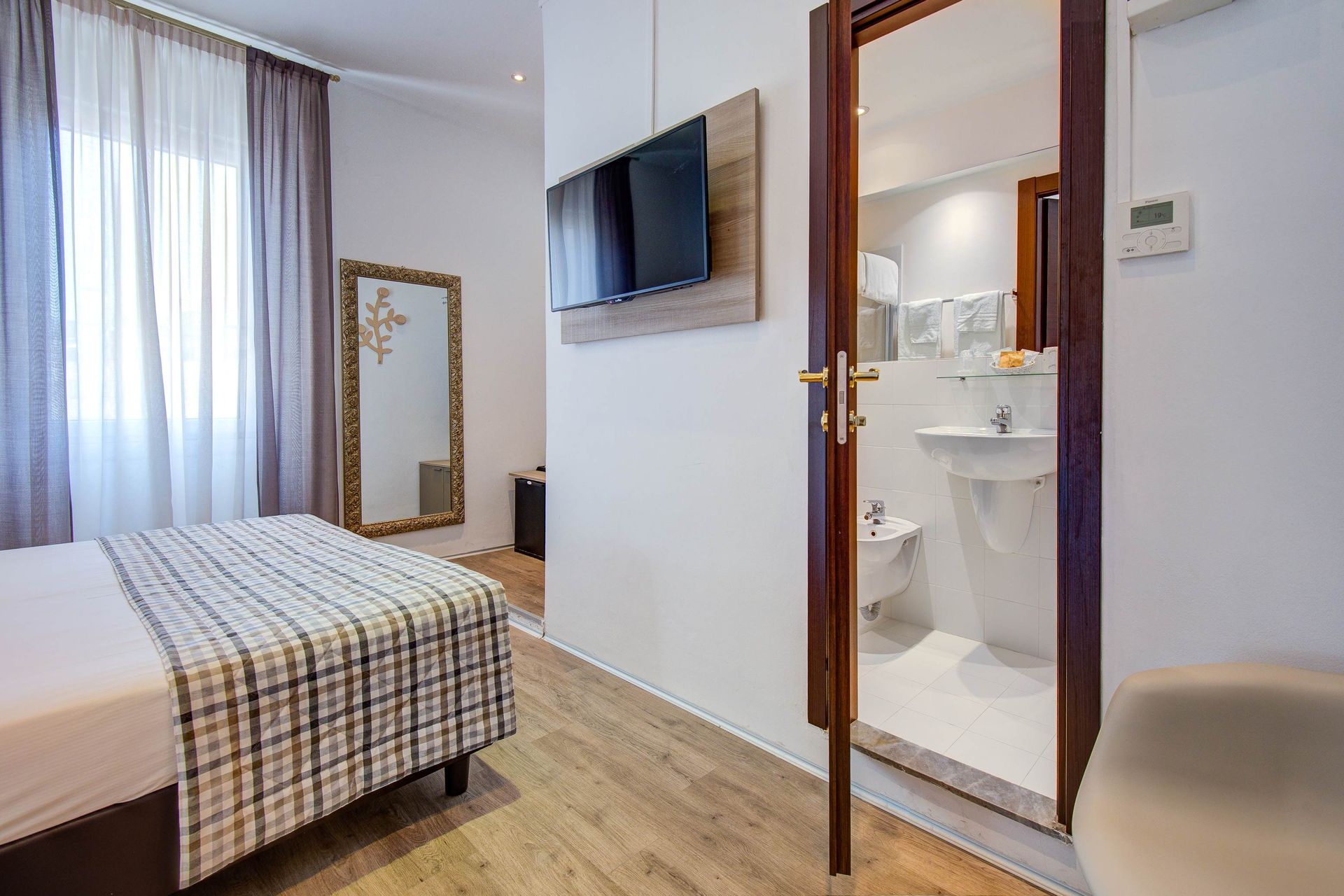 Interior de habitación de hotel con cama, TV, puerta del baño abierta, espejo y cortinas.