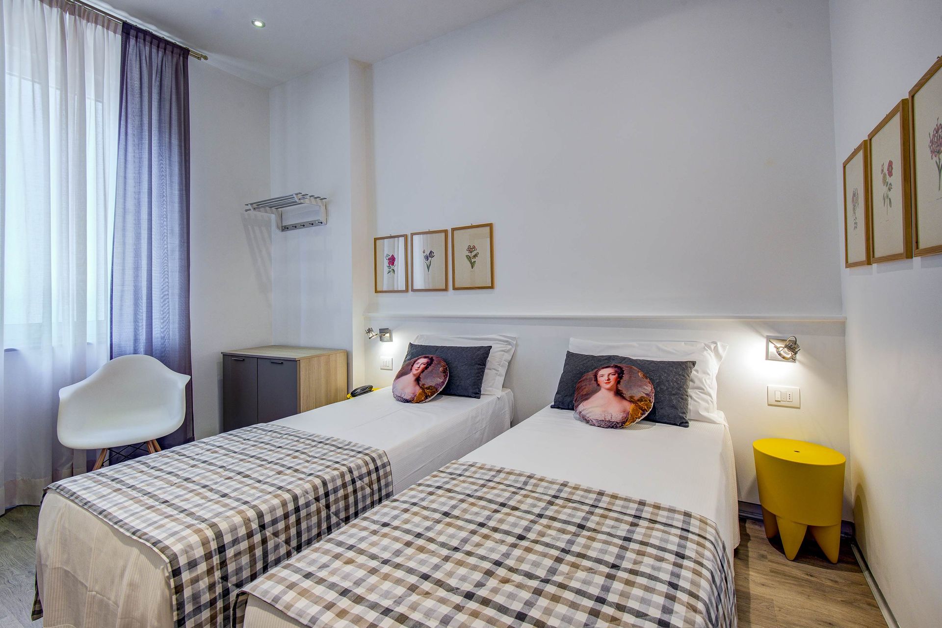 Camas individuales en una habitación bien iluminada con paredes blancas, arte enmarcado y una pequeña mesa amarilla.