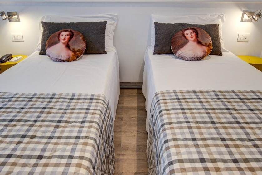 Dos camas individuales con sábanas blancas, mantas a cuadros y almohadas con retratos. Mesitas de noche amarillas.