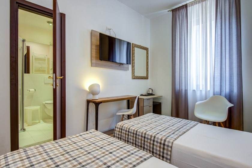 Habitación de hotel con dos camas individuales, pequeño escritorio, TV de pared y baño visible.