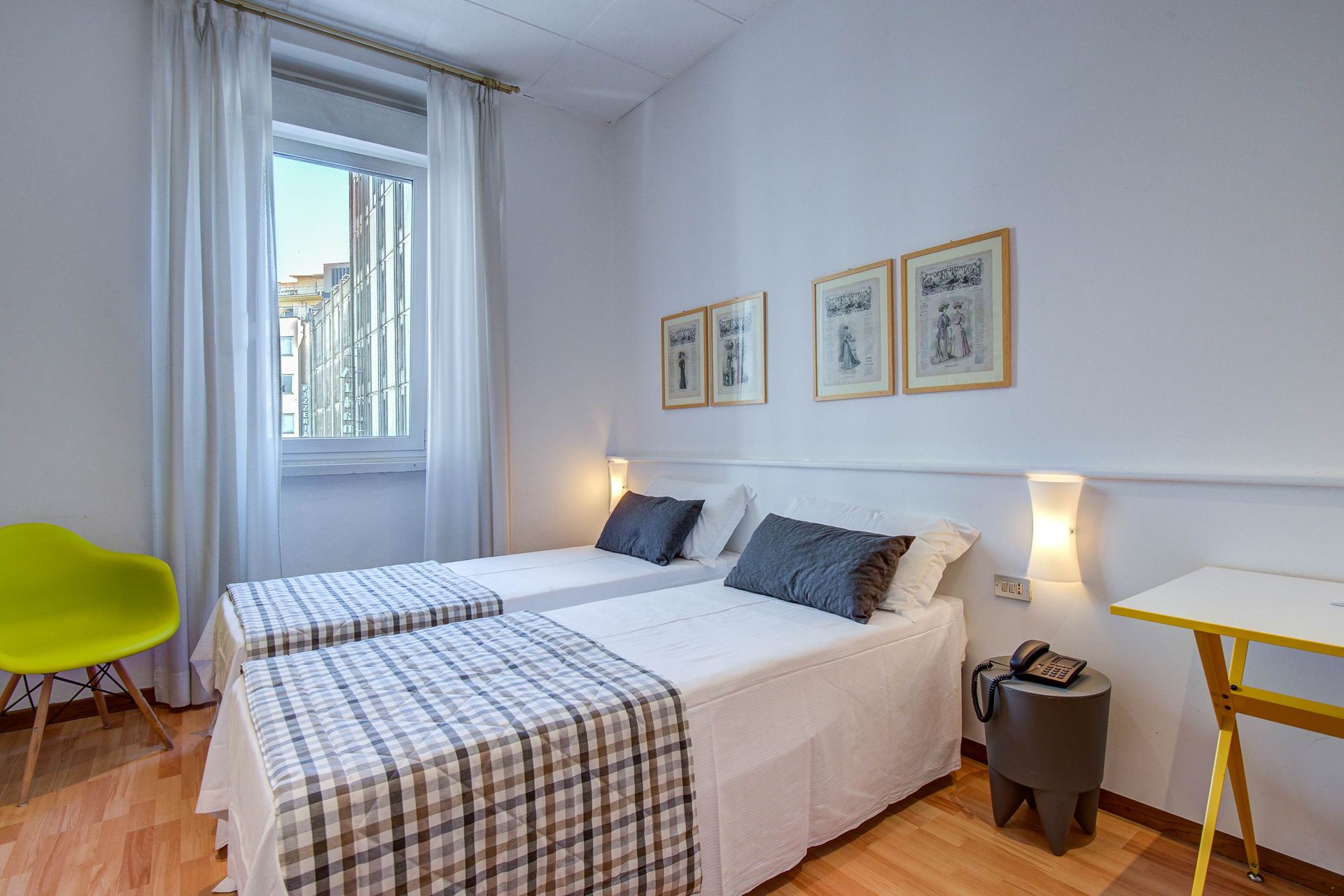 Habitación de hotel con dos camas individuales, paredes blancas, ventana, mantas a cuadros.