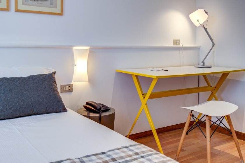 Dormitorio con cama blanca, escritorio amarillo, silla blanca, lámpara y piso de madera neutra.