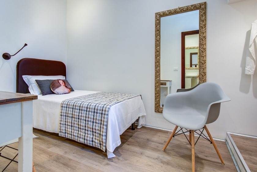 Dormitorio con una cama individual, espejo de cuerpo entero, silla moderna y un pequeño escritorio contra una pared blanca.