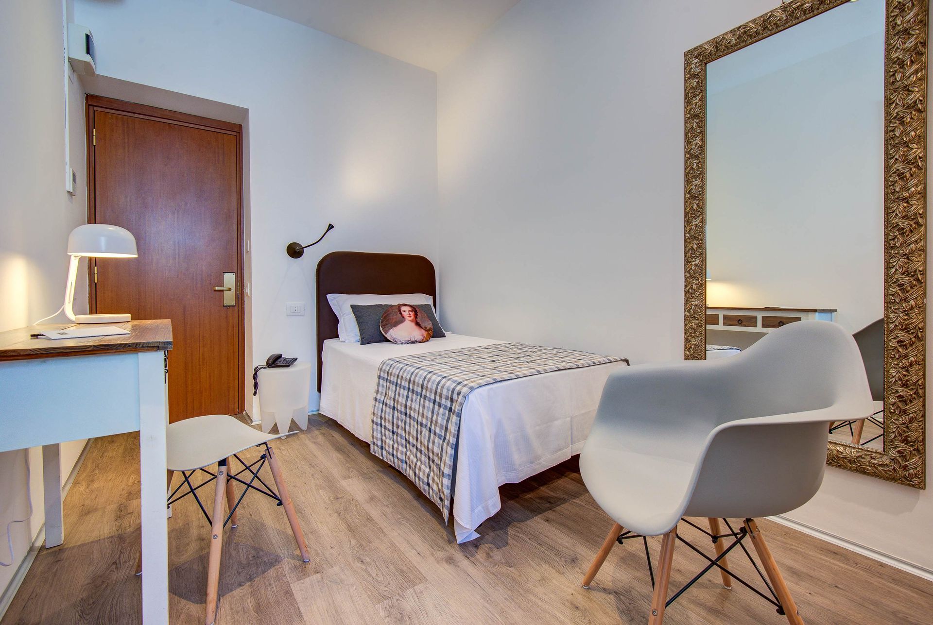 Dormitorio pequeño con cama individual, escritorio y espejo grande. Paredes blancas y suelo de madera.