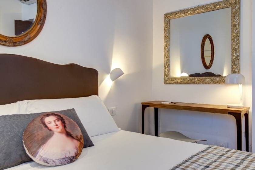 Dormitorio con cama, espejos y cojín con retrato. Cabecero marrón, paredes blancas y consola de madera.