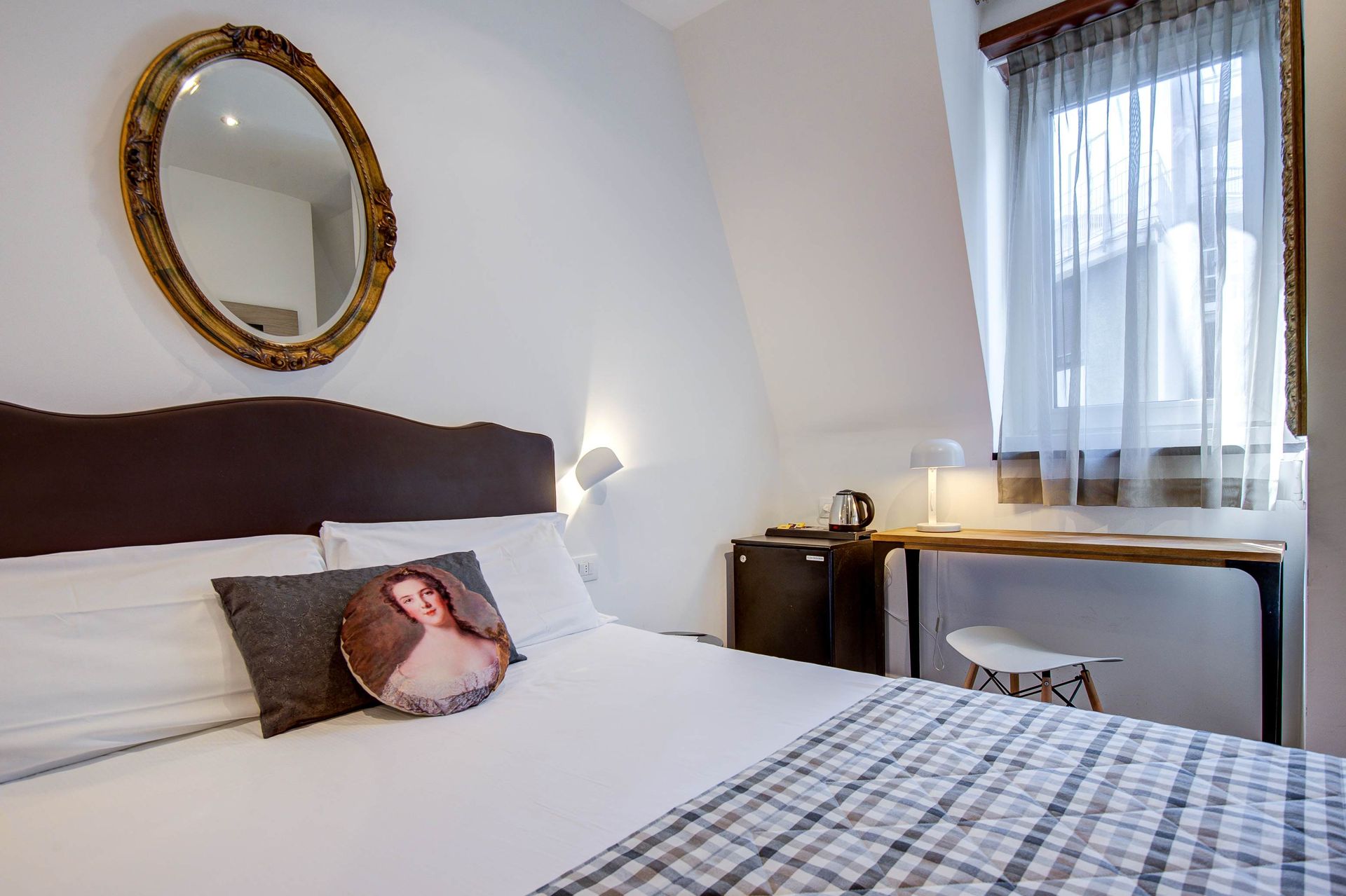 Dormitorio con cama, espejo, escritorio y ventana. Paredes blancas, marco de espejo dorado y manta gris y blanca.