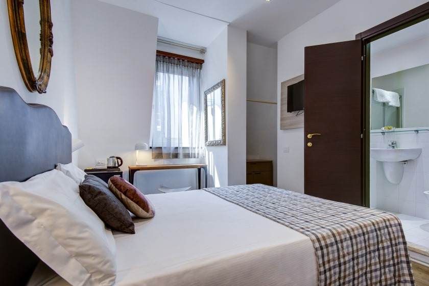 Dormitorio con cama, escritorio y baño privado. Paredes blancas, puerta marrón e iluminación tenue.