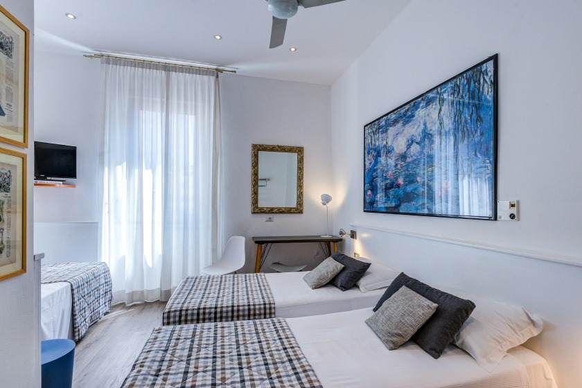 Habitación de hotel luminosa con tres camas, ventana, espejo y obras de arte. Paredes blancas y suelo de madera clara.