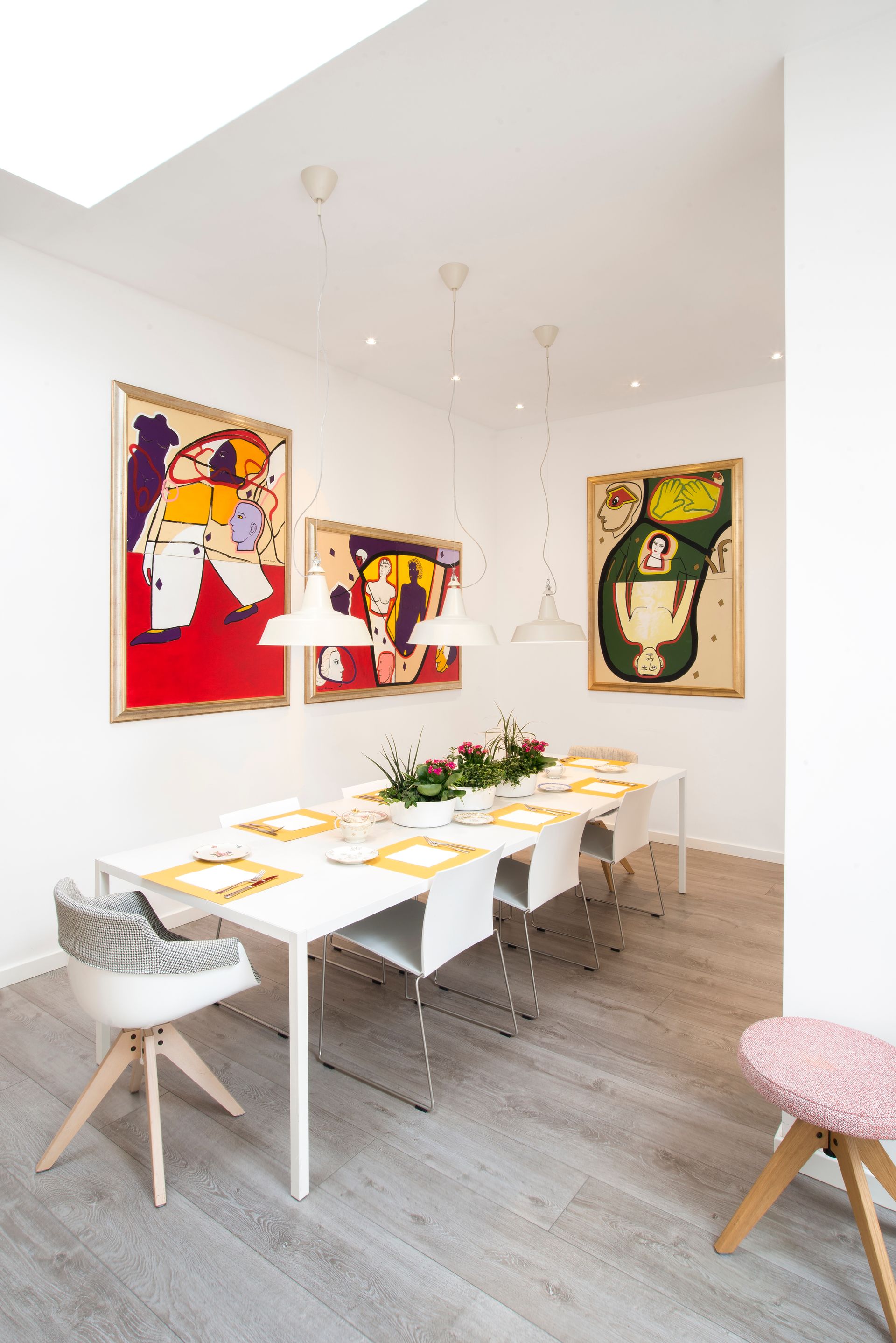 Comedor con arte abstracto, mesa blanca puesta para invitados, sillas modernas y lámparas colgantes.