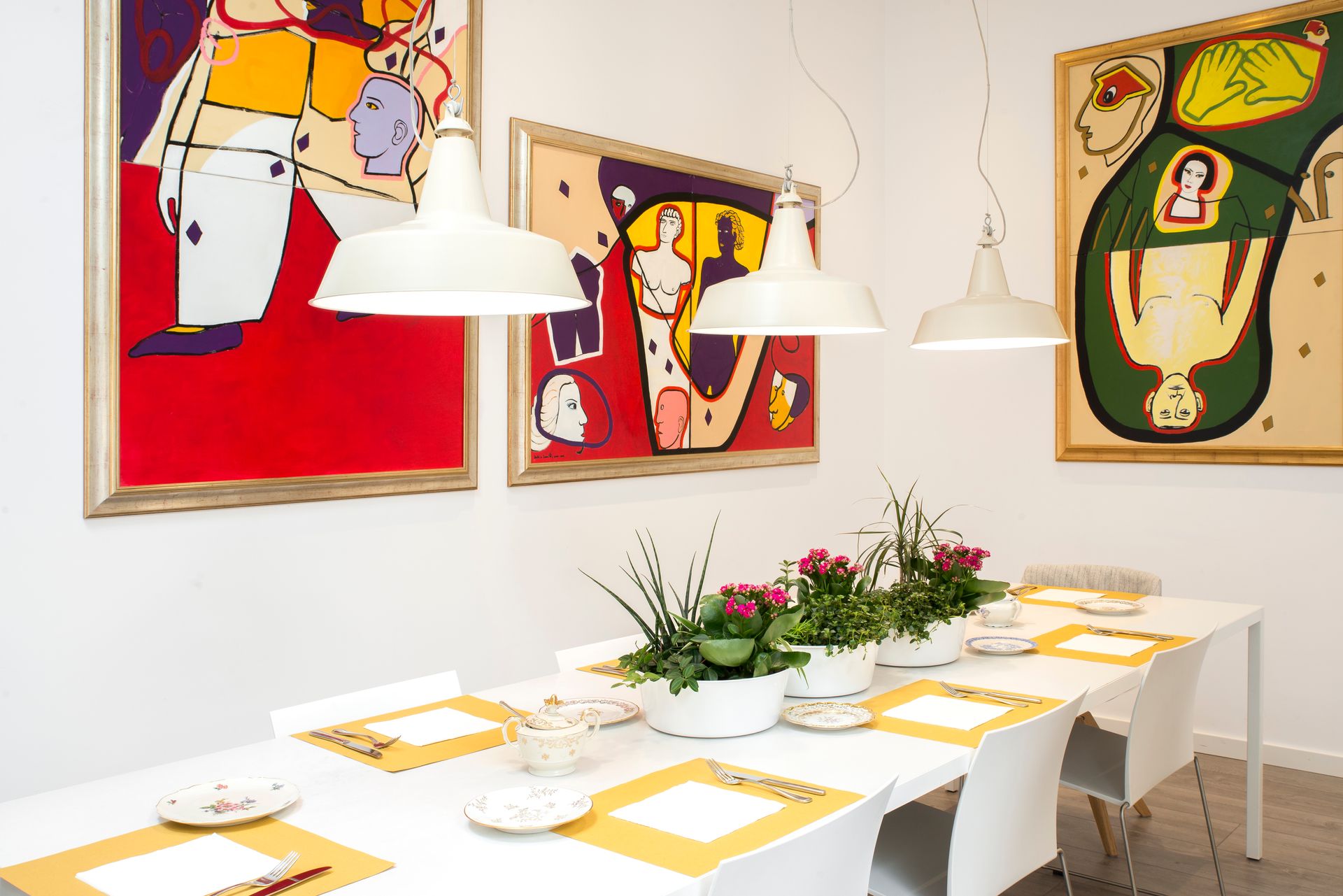 Comedor con arte en paredes blancas, mesa larga con cubiertos y lámparas colgantes.