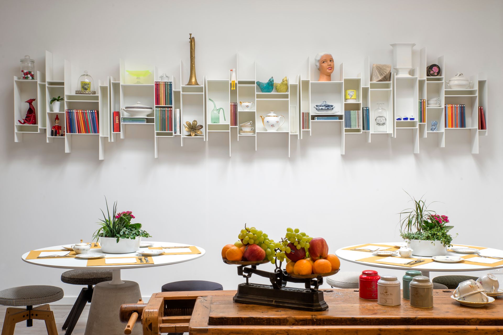 Estantería blanca sobre dos mesas redondas, con libros y decoración, y una balanza de frutas sobre una mesa de madera.