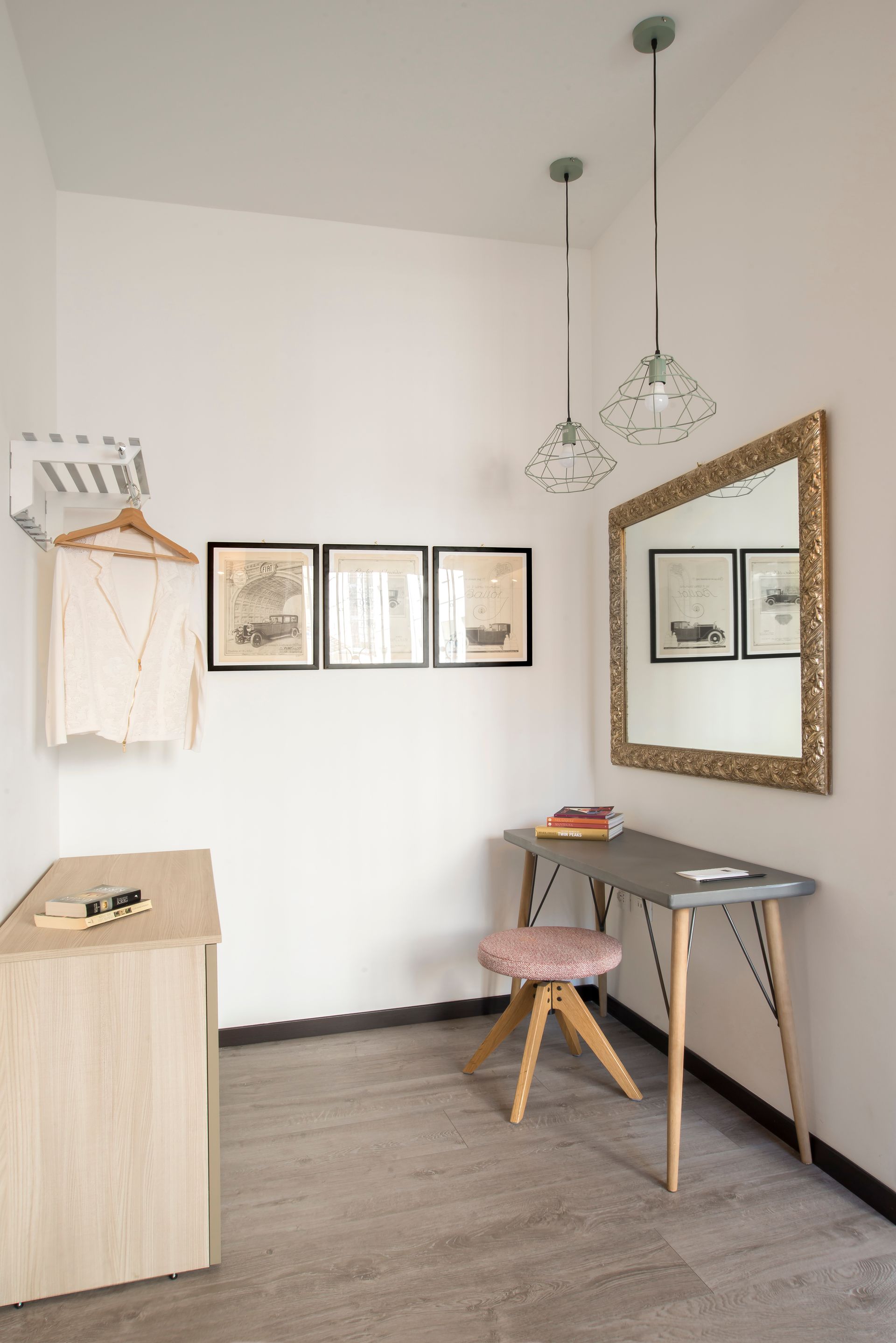 Una habitación pequeña y luminosa con un escritorio, un espejo, obras de arte y un perchero con una camisa.