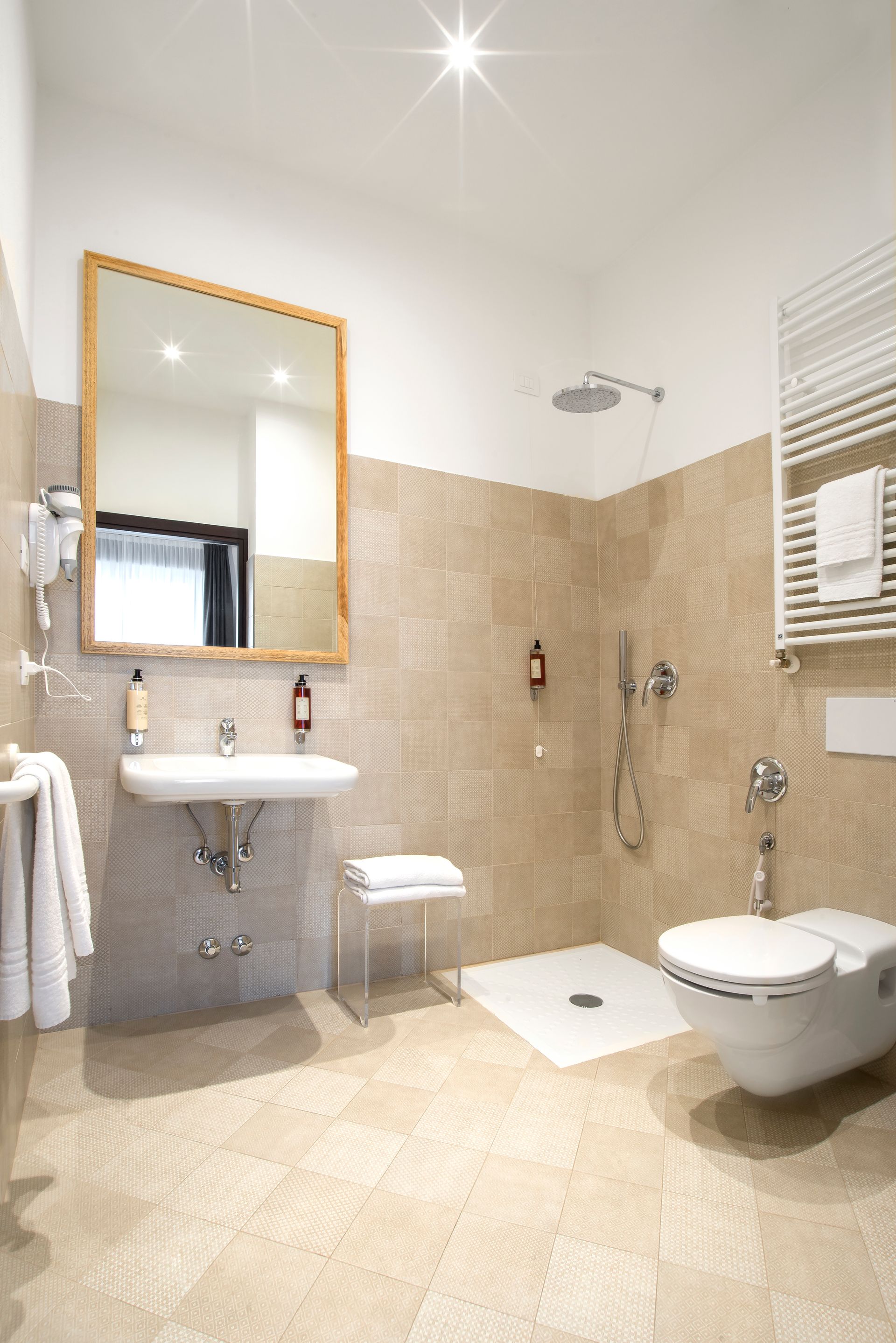 Baño luminoso con espejo cuadrado, lavabo, ducha e inodoro. Azulejos claros cubren las paredes y el suelo.