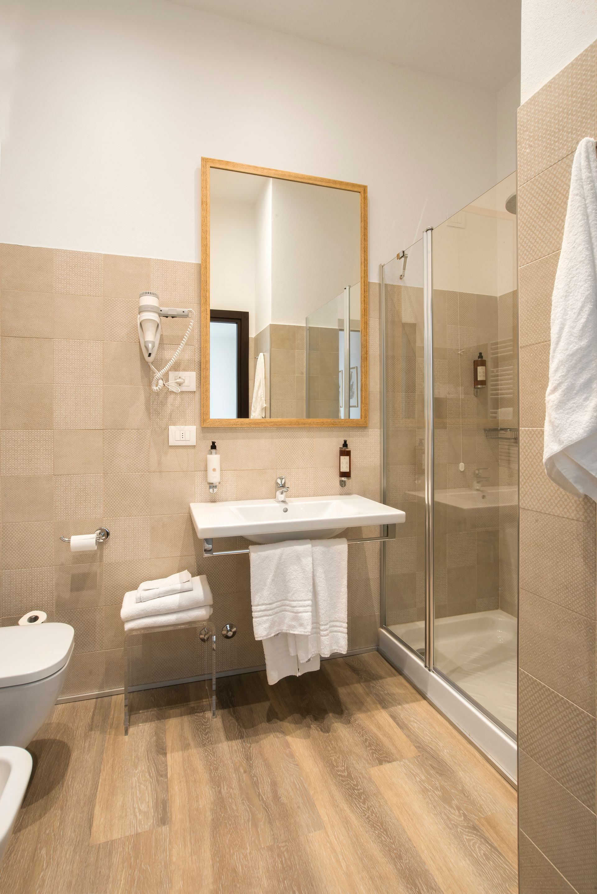 Baño con paredes beige, suelo de madera, ducha de cristal, lavabo blanco y espejo grande.