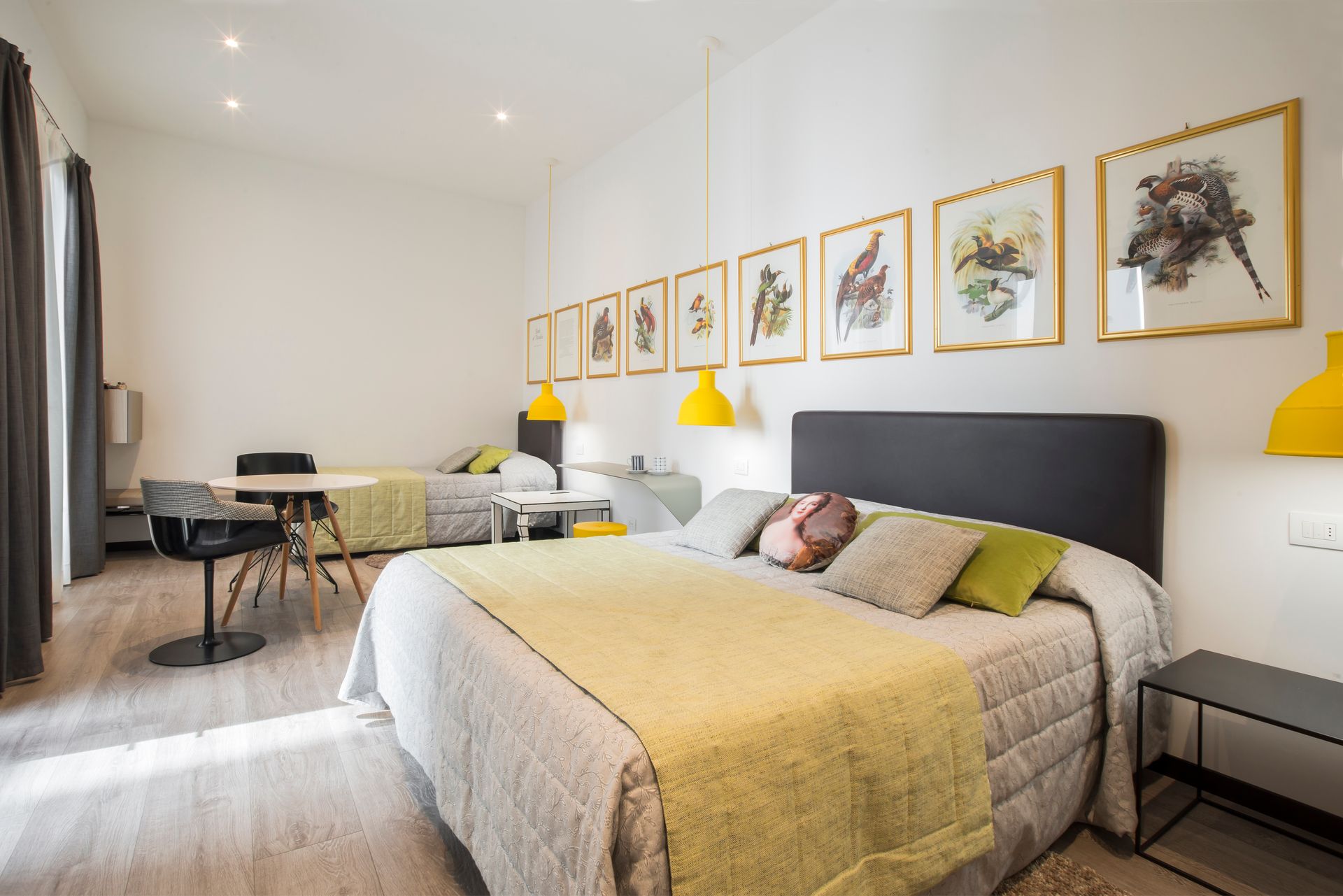 Dormitorio con una cama grande, una cama pequeña, una mesa con sillas, lámparas colgantes amarillas y arte enmarcado.