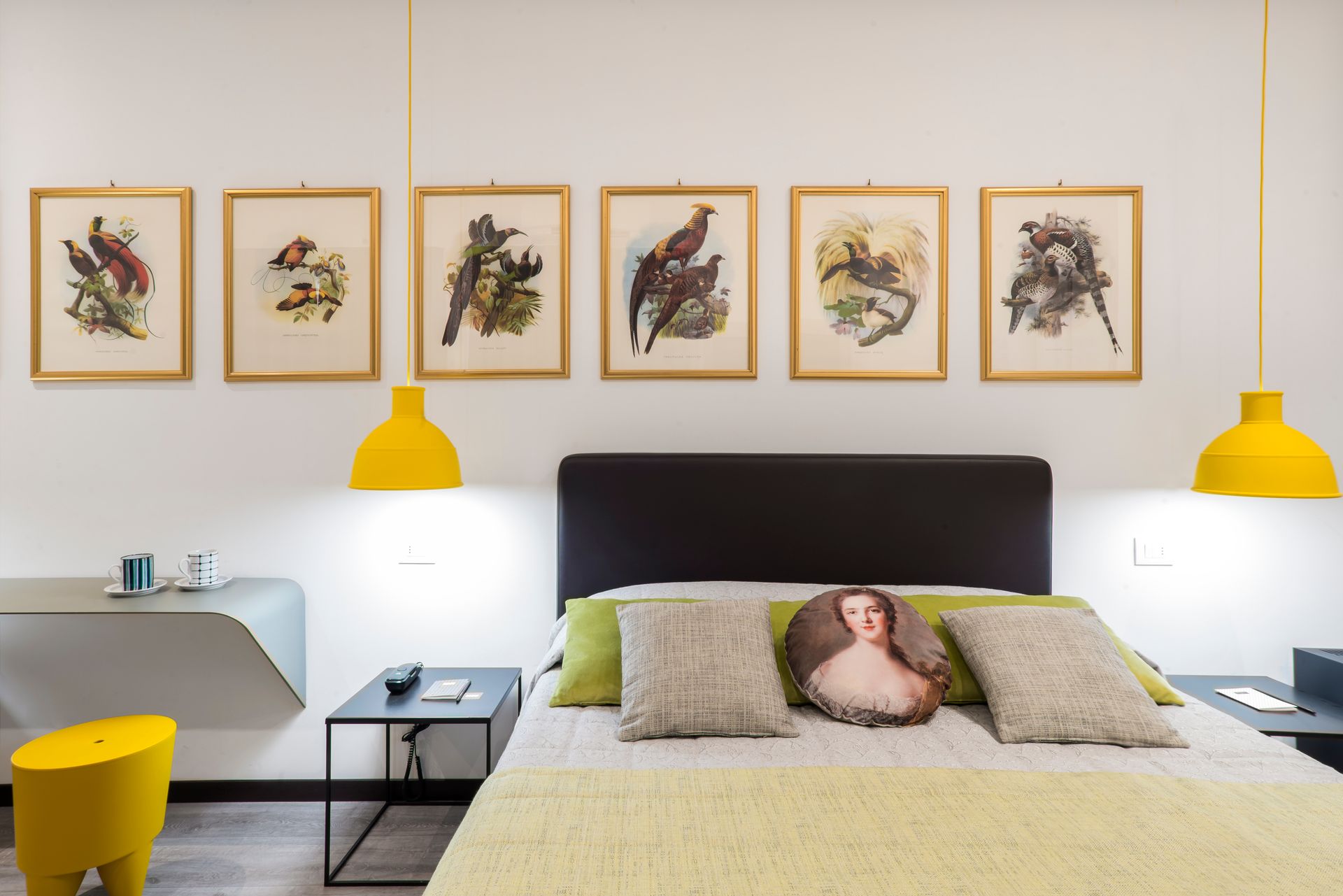 Dormitorio con arte de pájaros, lámparas amarillas, cama con almohadas y una mesa auxiliar gris.