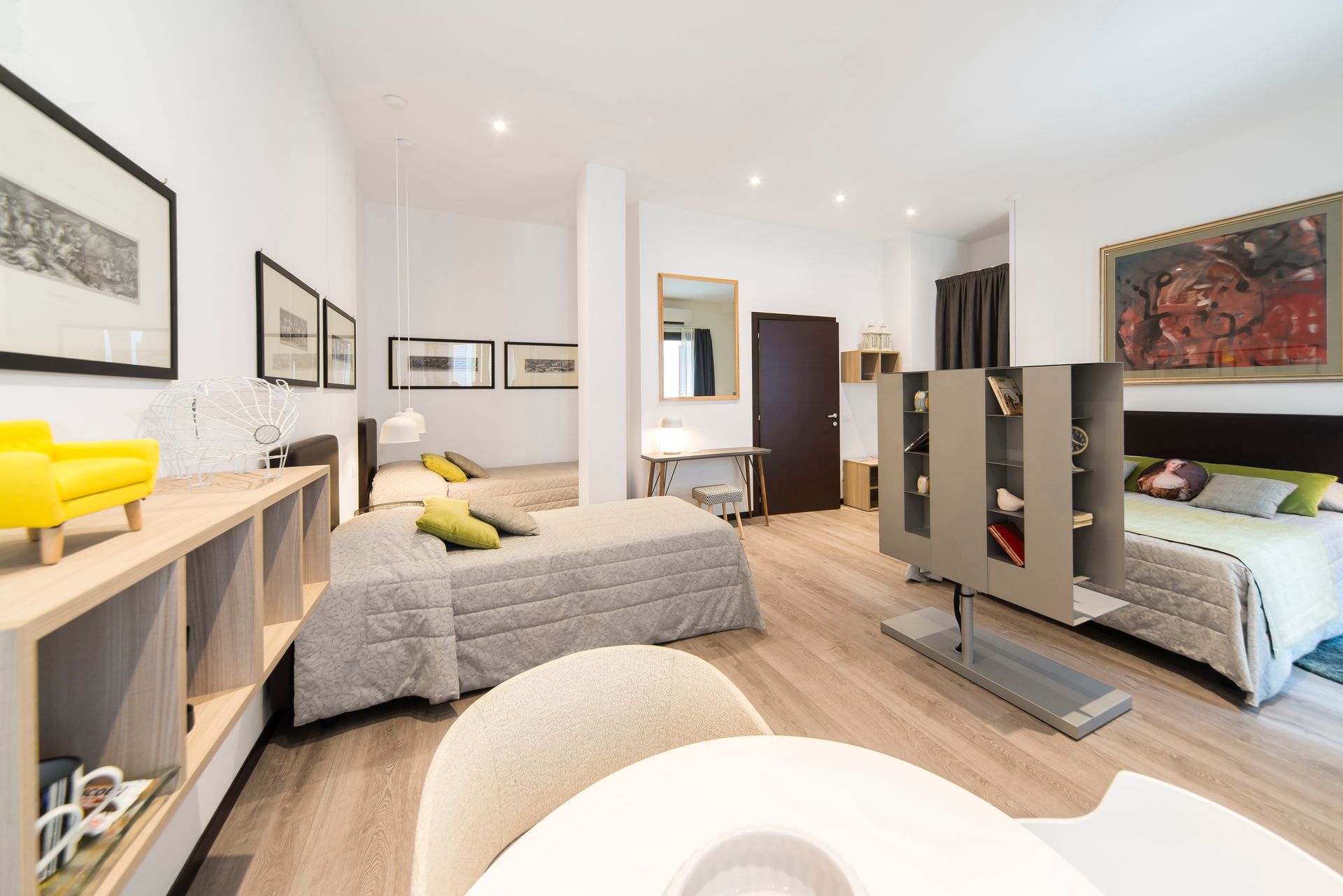 Habitación de hotel moderna y luminosa con tres camas, suelos de madera, obras de arte y un biombo parcial.