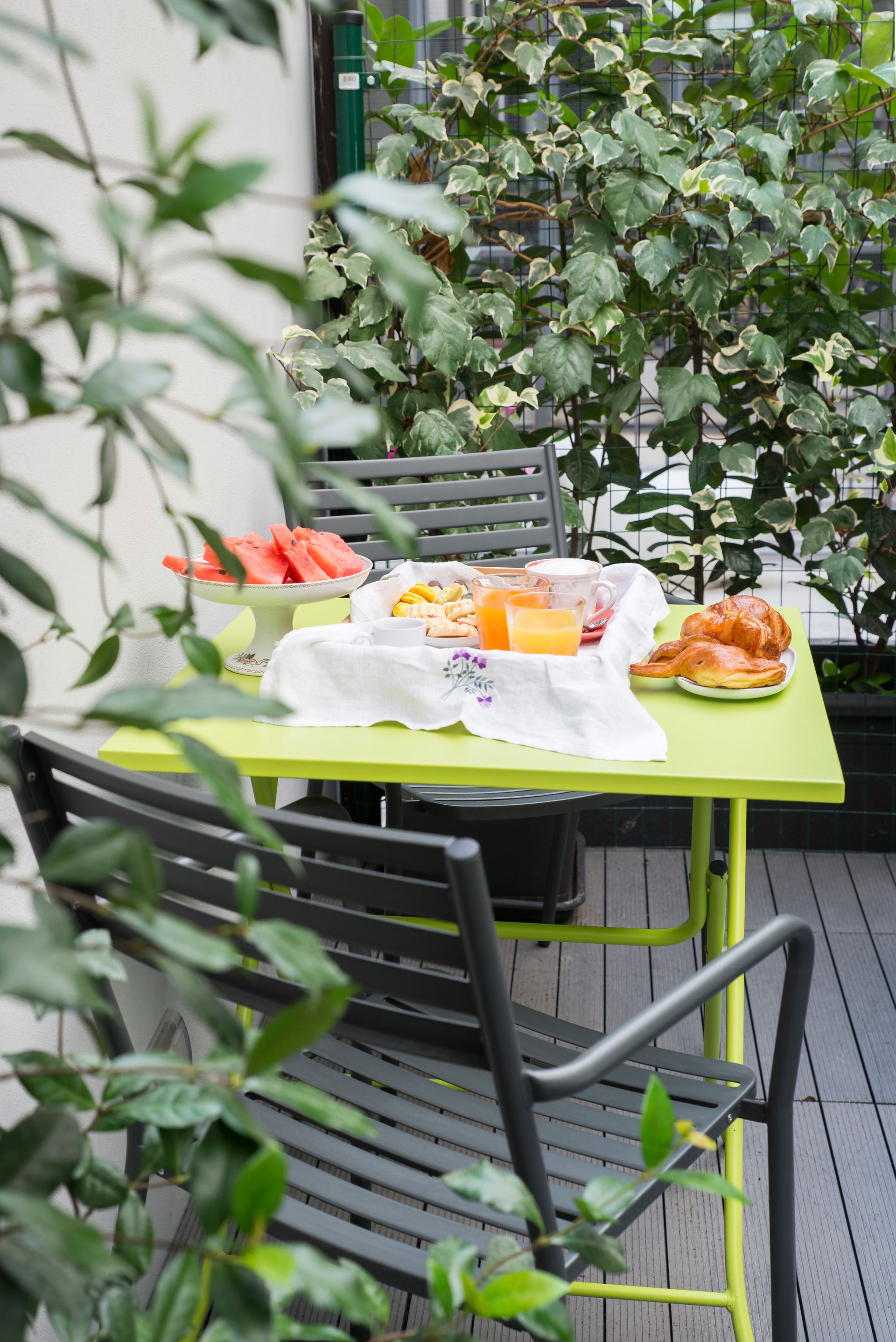 Mesa verde lima dispuesta para desayunar con sandía y croissants en un balcón con plantas.