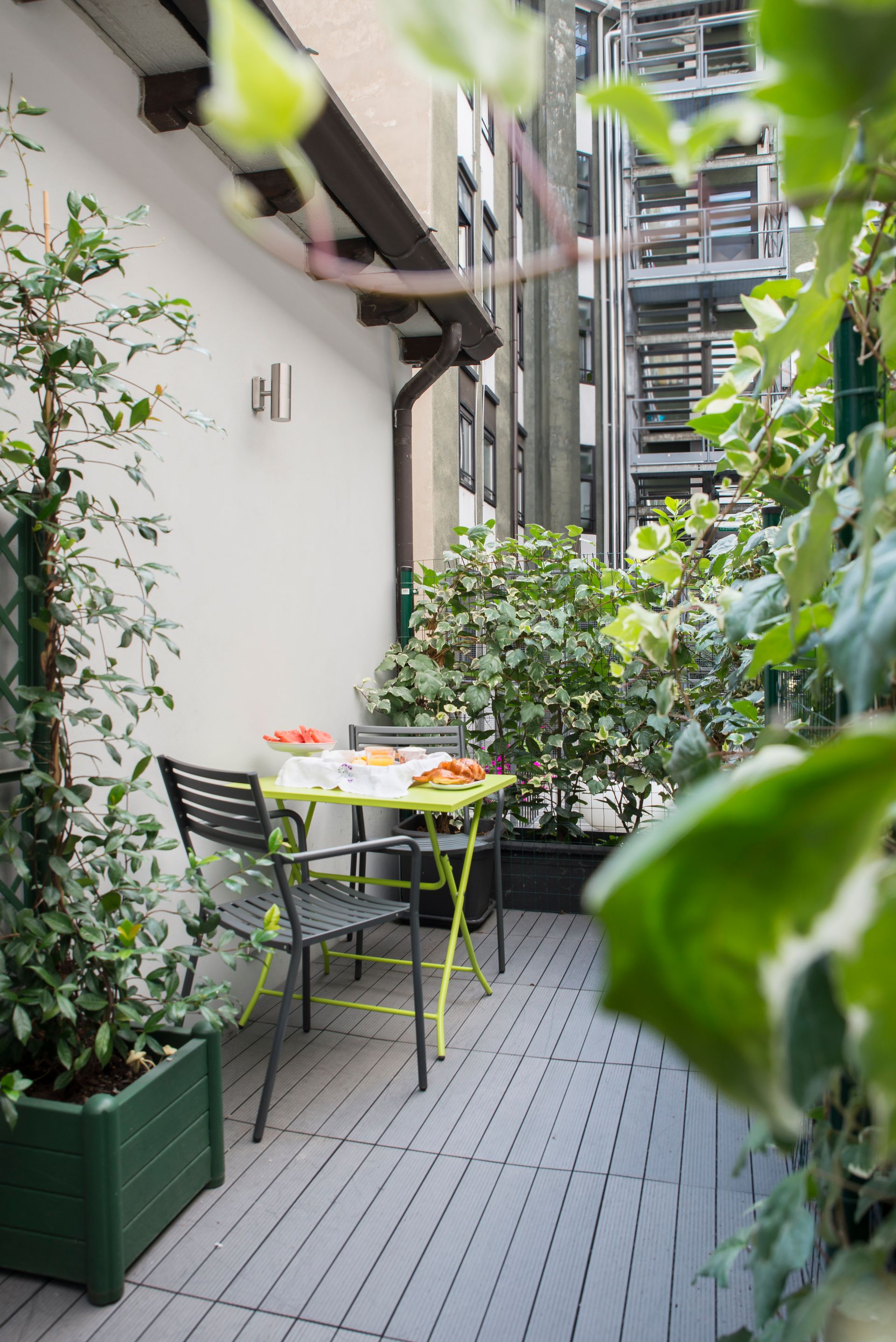 Pequeño balcón con mesa y sillas rodeado de vegetación, en un entorno urbano.
