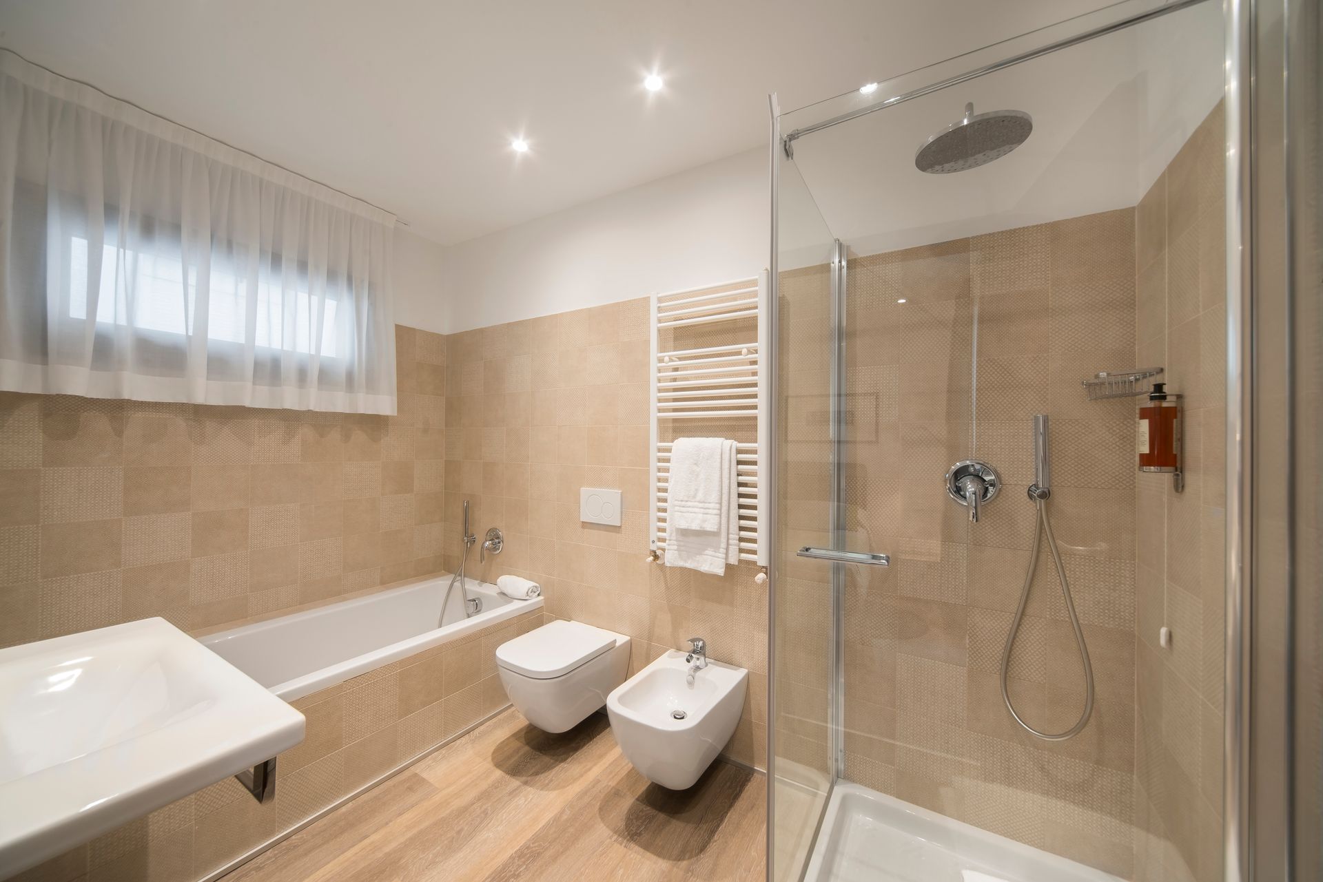 Baño con bañera, ducha, inodoro y bidé. Paredes beige, suelo imitación madera clara y grifería blanca.