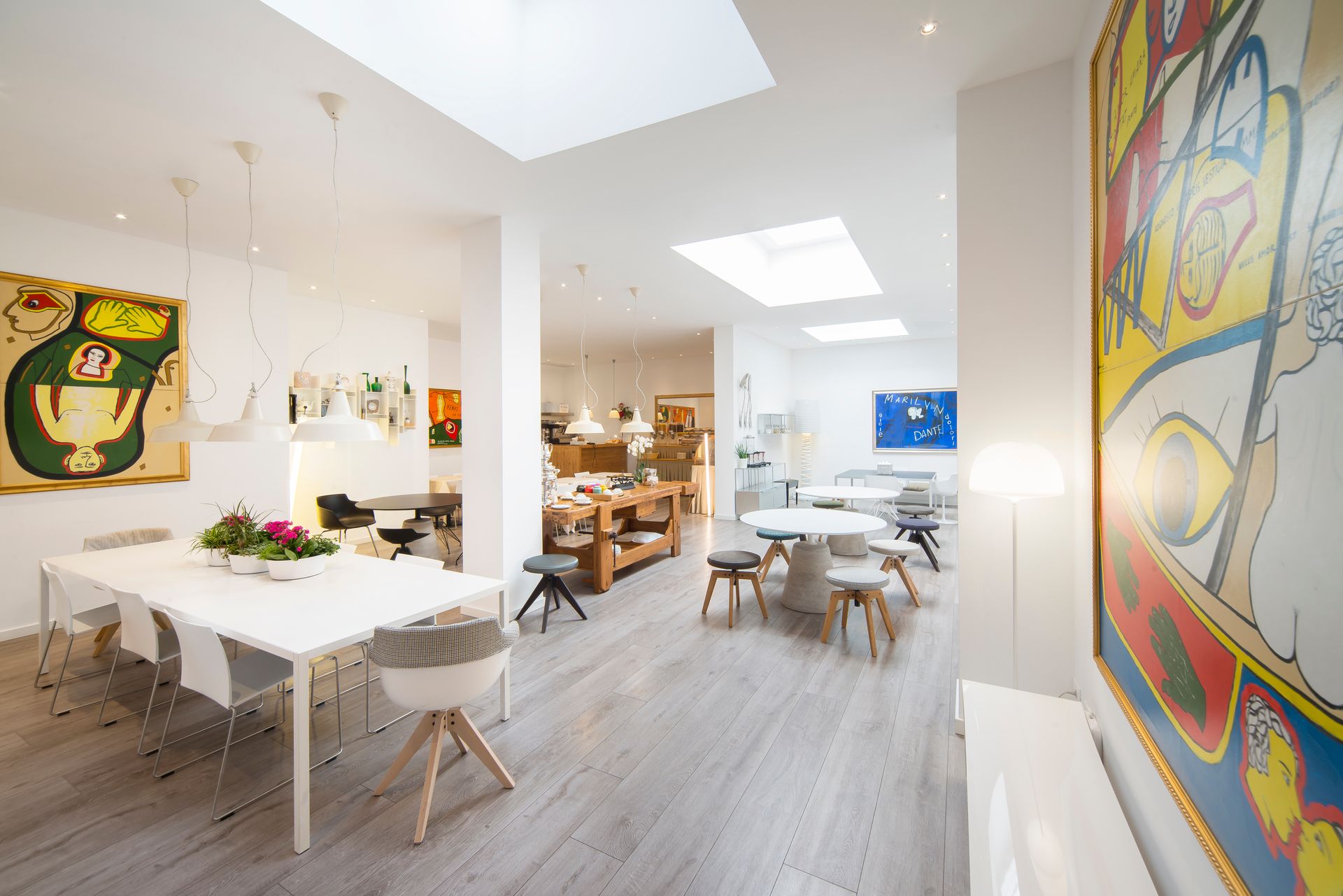 Interior de galería de arte moderno y luminoso con paredes blancas, tragaluces y obras de arte, con mesas y sillas.