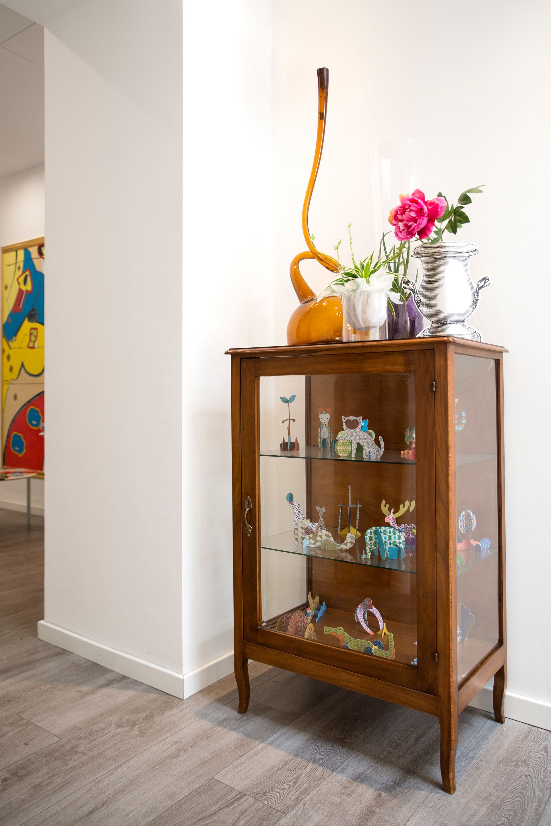 Vitrina de madera con puerta de cristal. Flores, objetos de cristal y un instrumento musical encima.