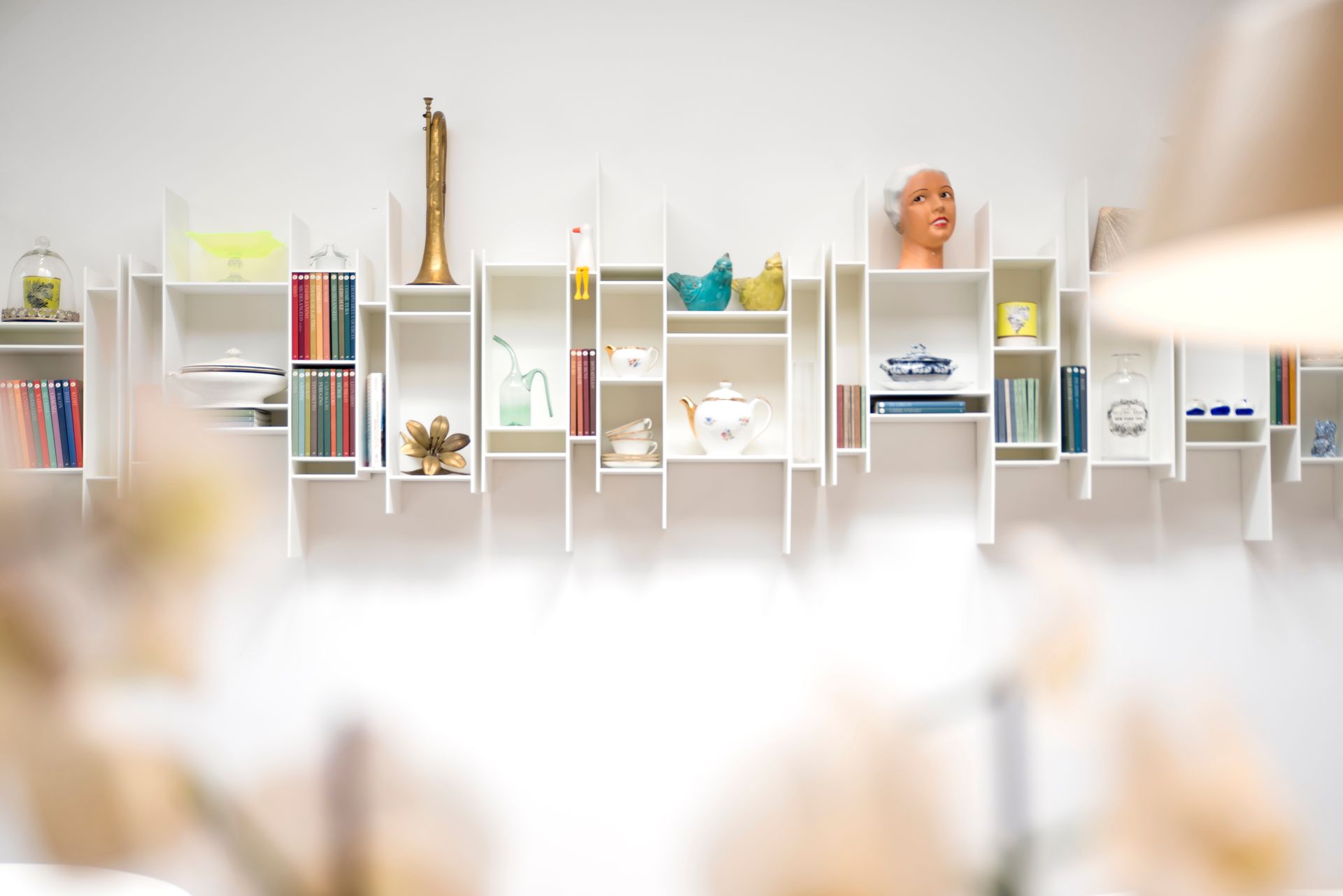 Estantes modulares blancos en una pared exhiben diversos objetos, incluidos libros, figuras y una cabeza de maniquí.