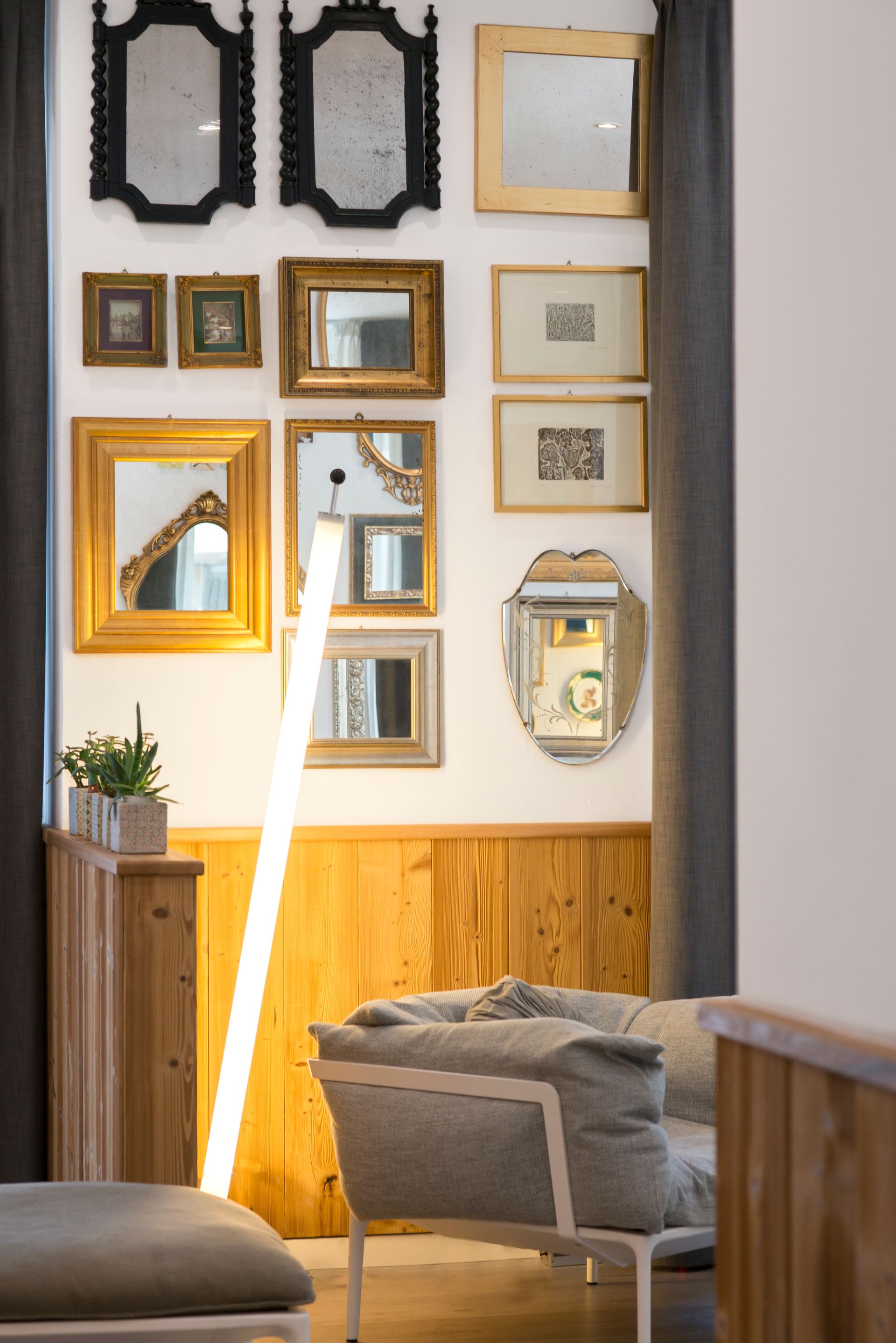 Pared de galería con espejos y arte enmarcado, con una lámpara de pie moderna, paneles de madera y un sofá.