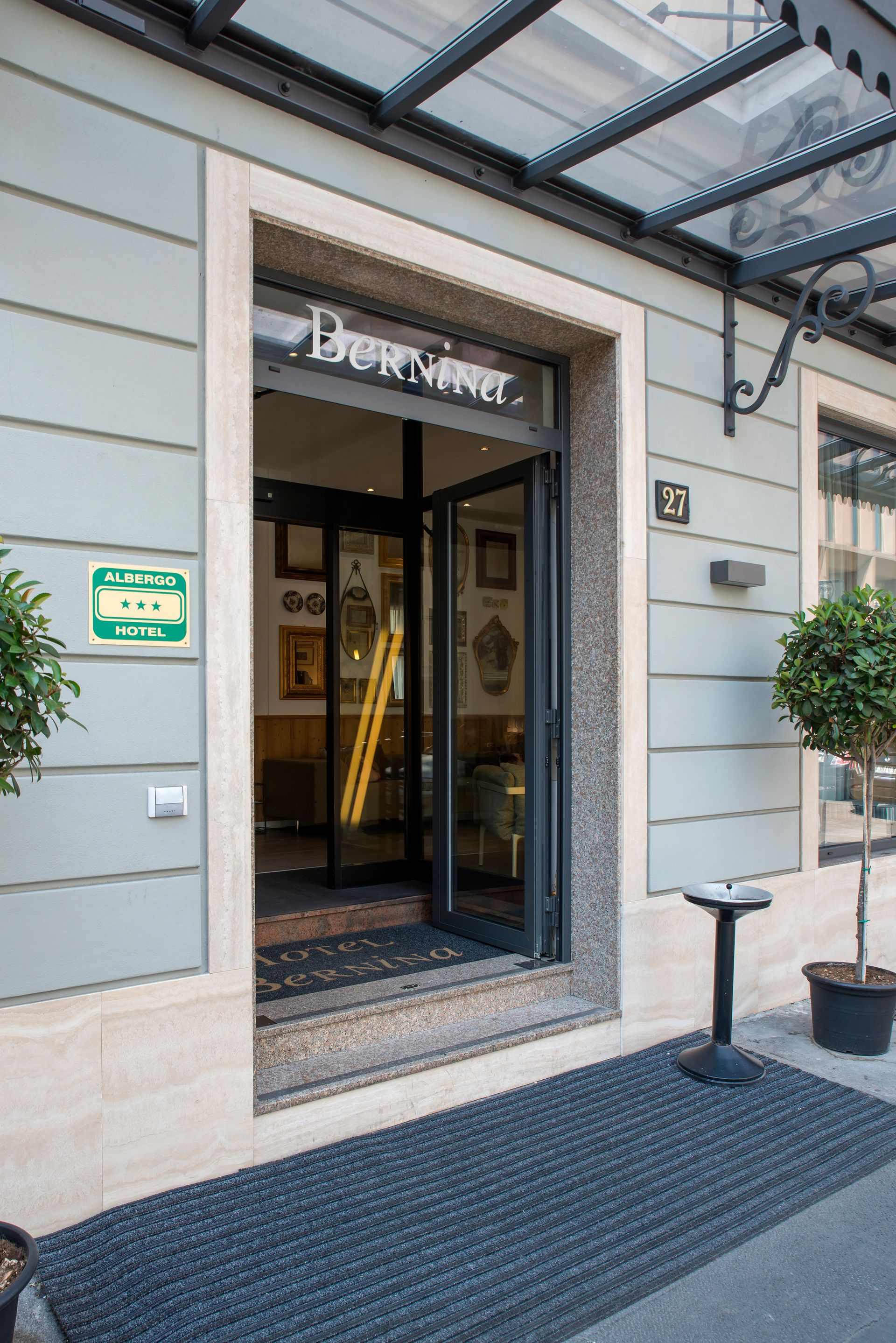 Entrada a un edificio con puertas de cristal, enmarcado por piedra gris. Se ven un letrero verde y plantas en macetas.