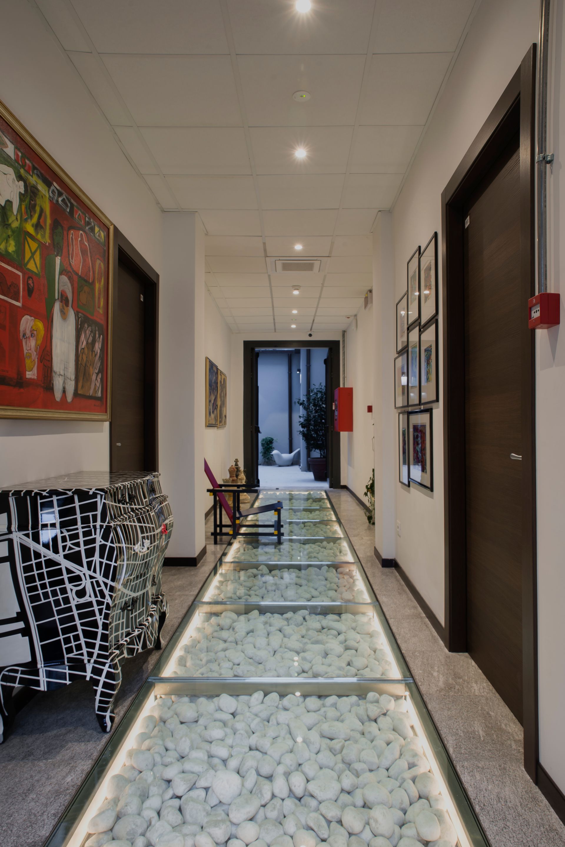 Pasillo largo con piso de vidrio sobre piedras blancas, puertas oscuras, arte y un armario.