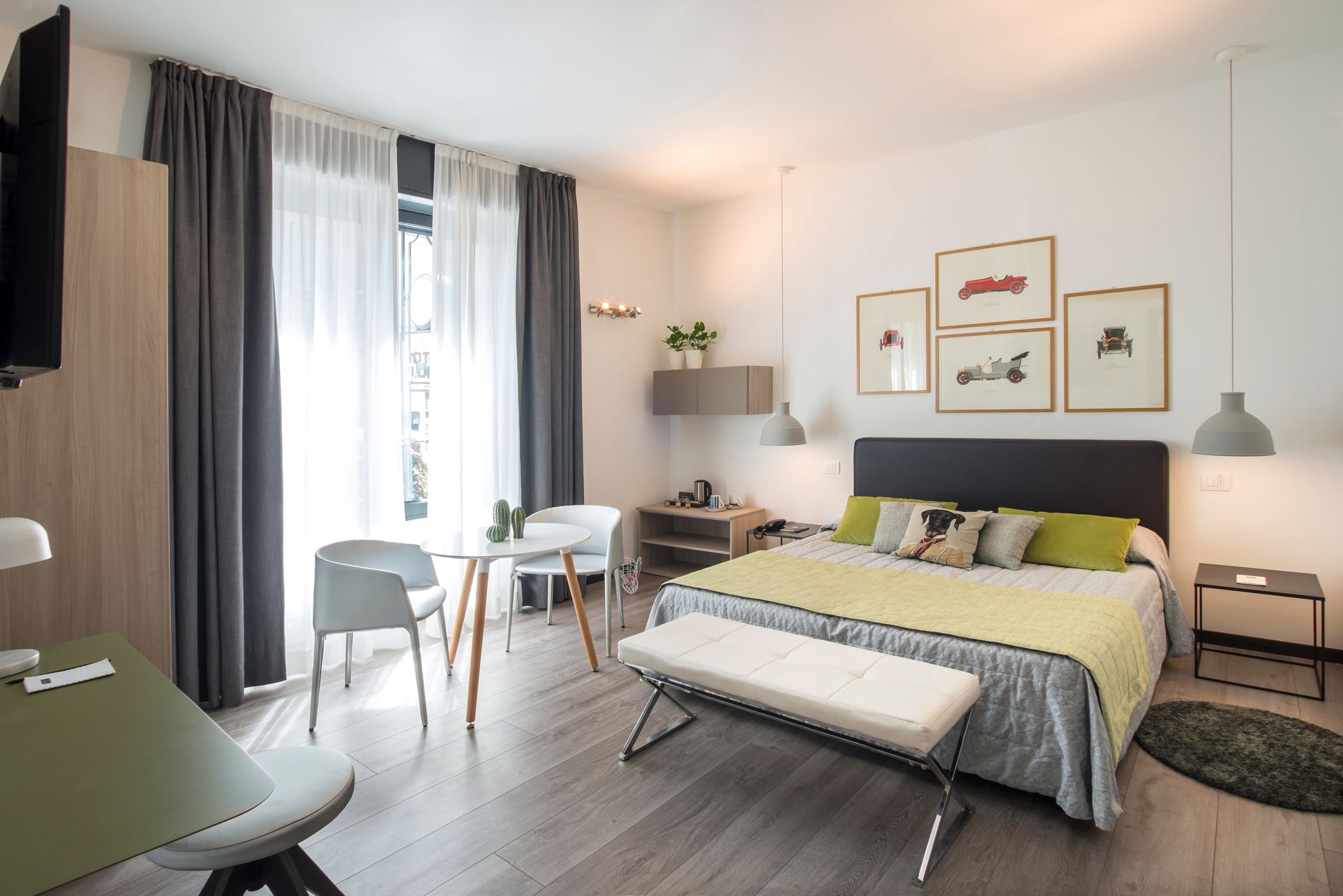 Bright hotel room with bed, table, chairs, art, gray curtains, and wood flooring.
