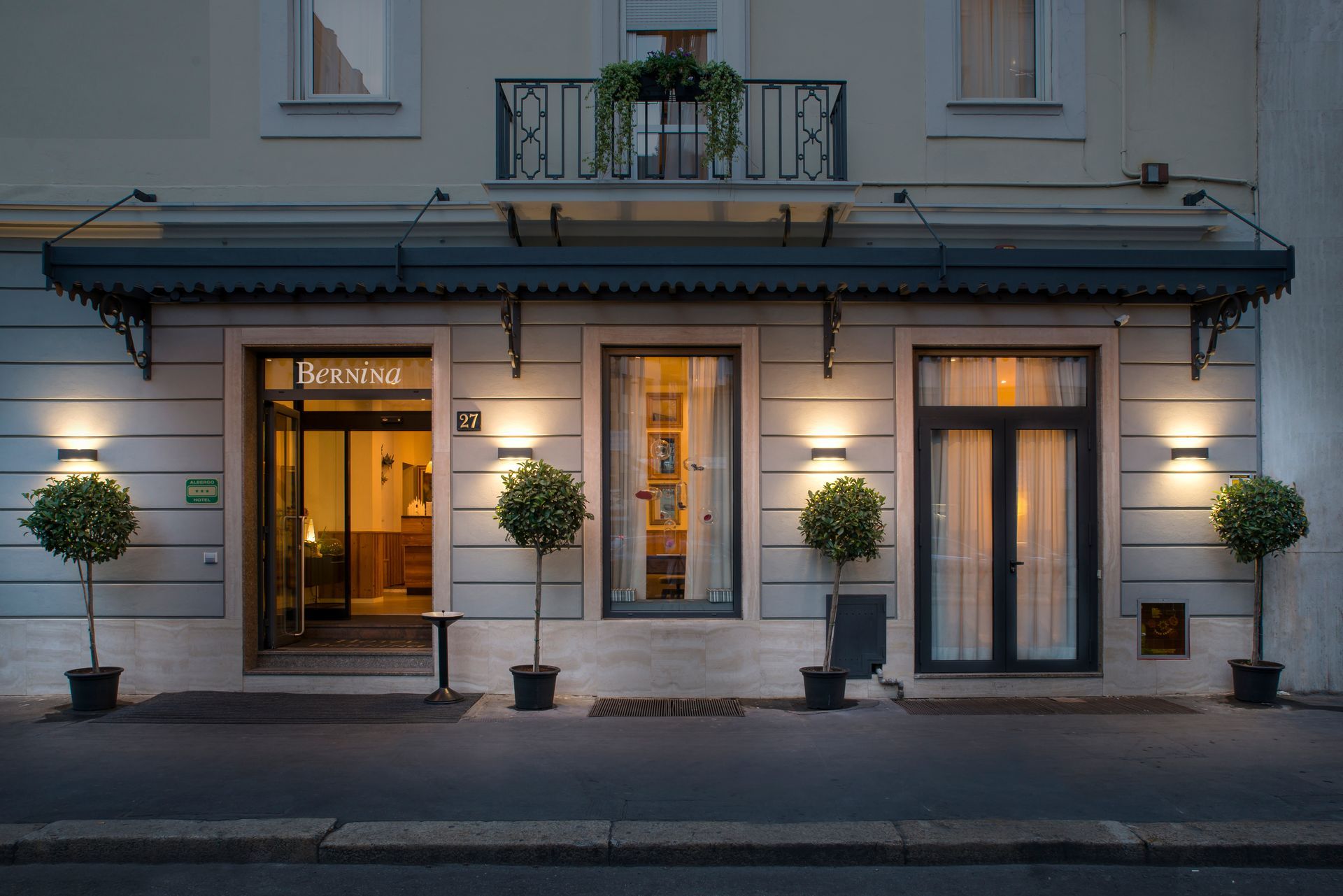 Hotel entrance with awning, lit by wall-mounted lights. Small trees flank the doors.