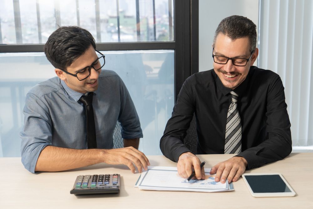 Two men reviewing documents at a desk, smiling. Calculator and tablet present, with a window in the background.