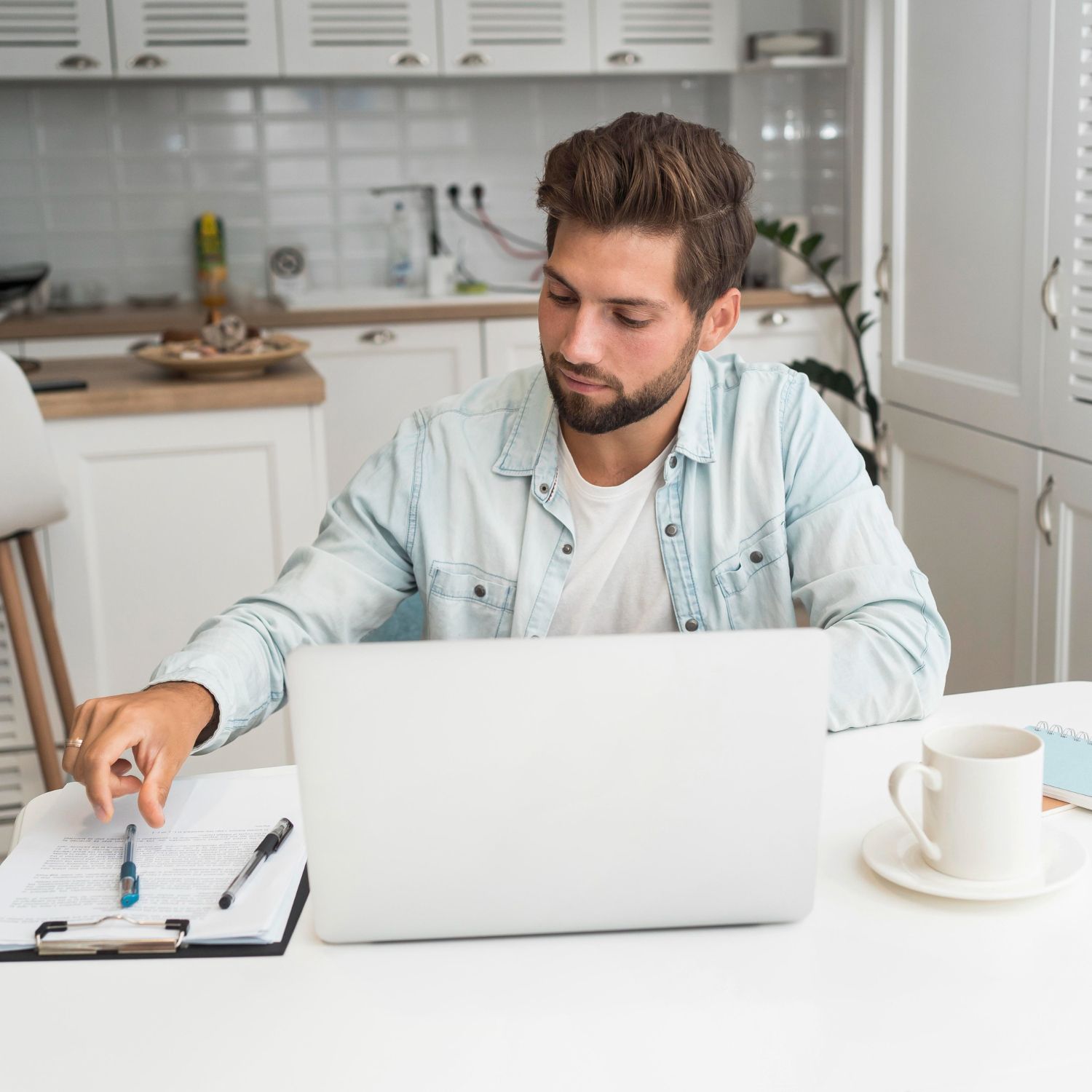 Man in denim shirt at a laptop, working at a kitchen table with documents and coffee nearby.