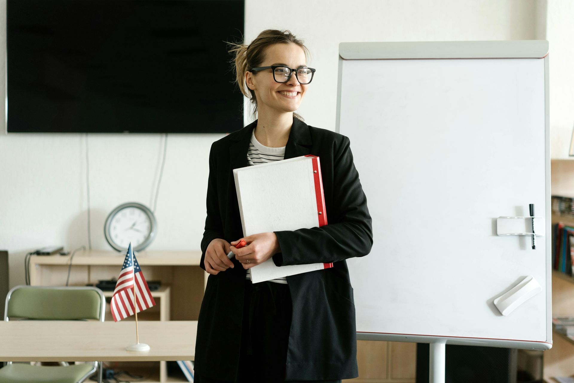 Woman wearing glasses and blazer, holding papers, standing by a whiteboard and an American flag in a classroom.