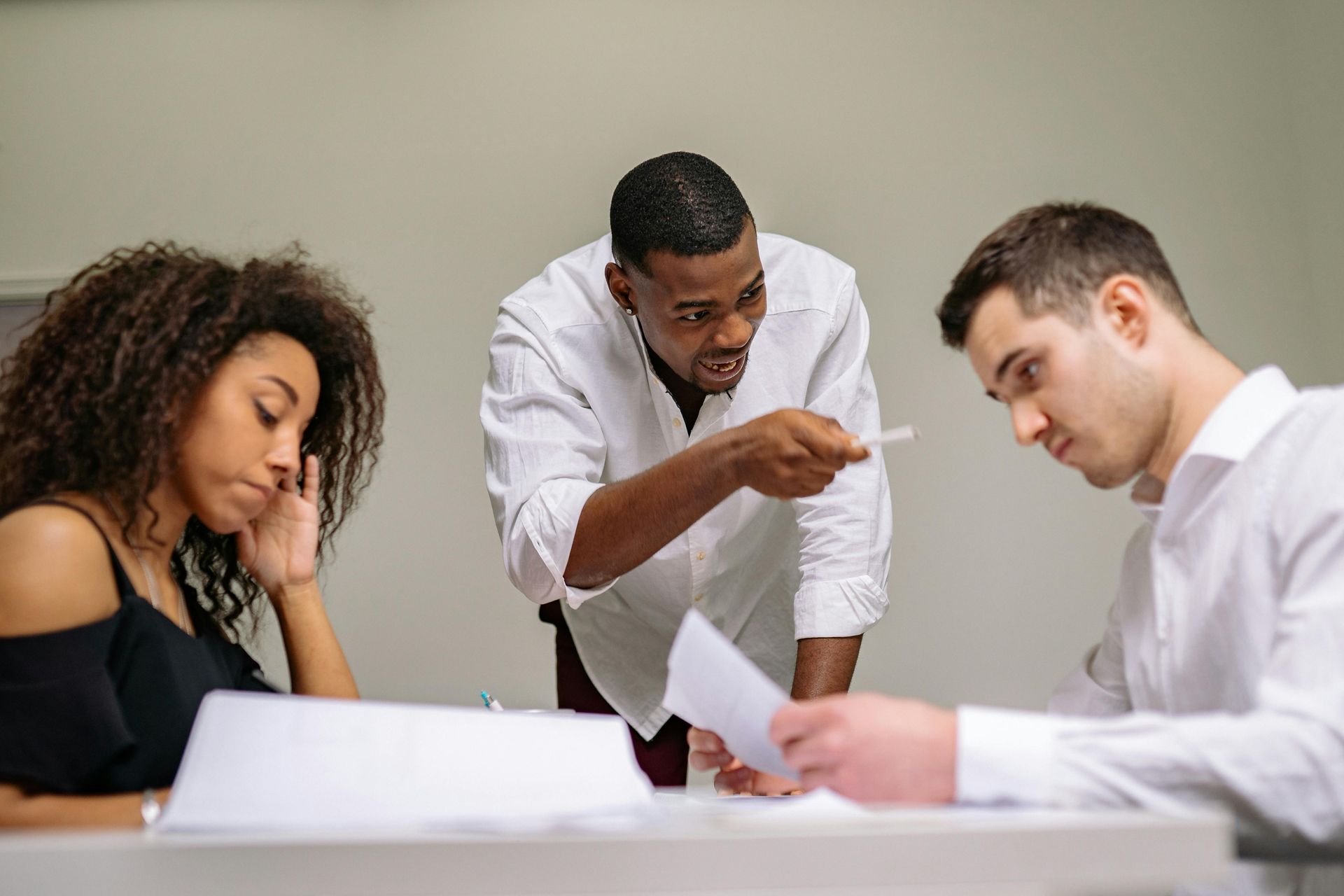 Man gestures angrily at paperwork while two others look on, one with a concerned expression.