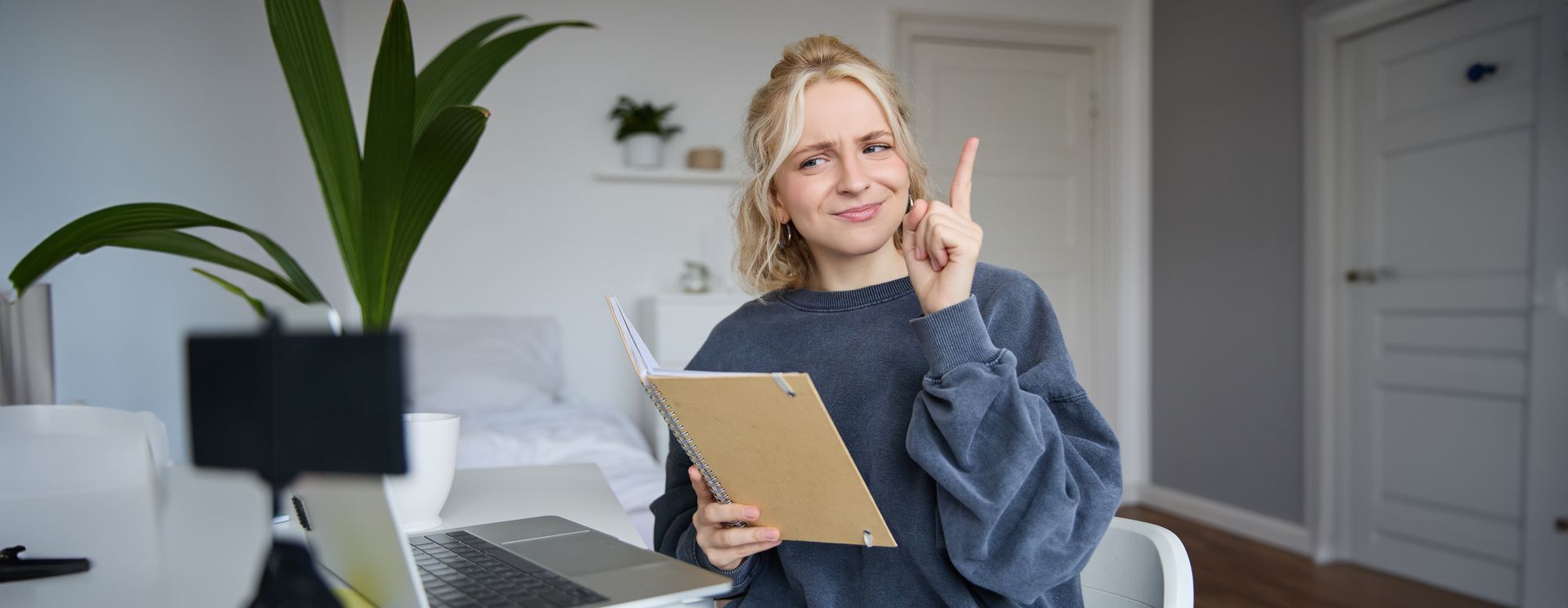 Woman holding notebook, pointing upwards, smiling, while sitting near a laptop, plant, and phone stand.