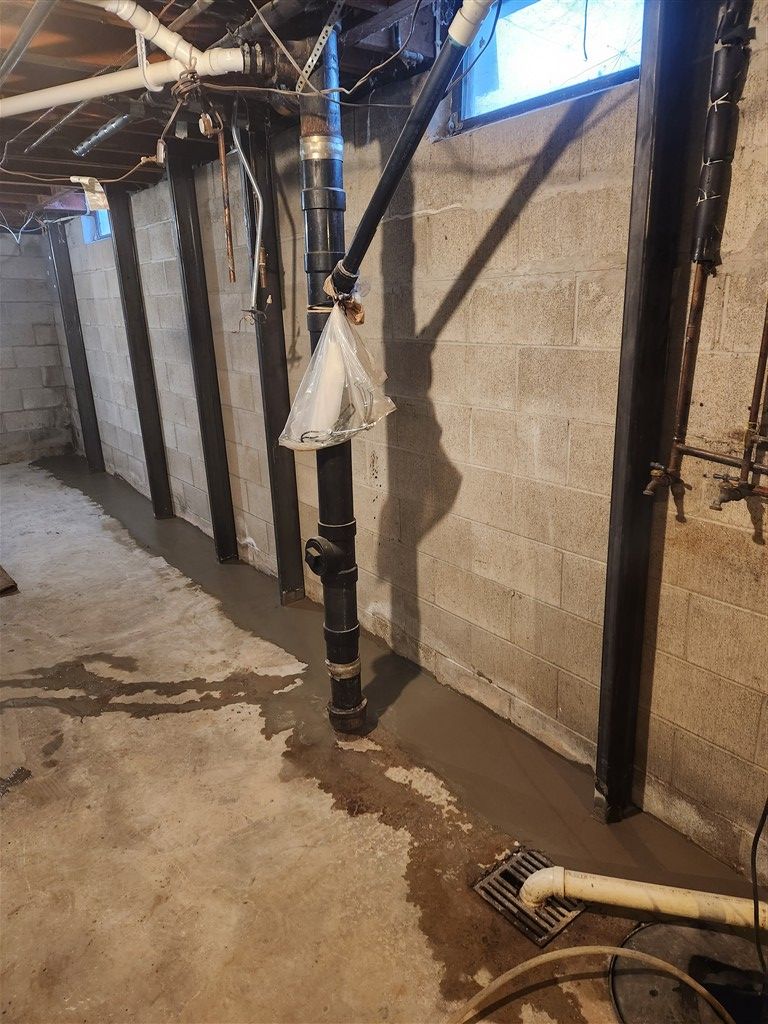 Basement with concrete walls and floor. Black pipes, drain, and light. 
