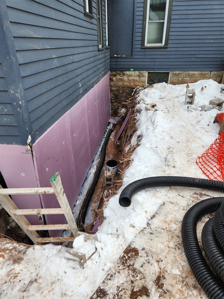 Excavation alongside a house. Pink insulation is installed, a black drainpipe is visible.