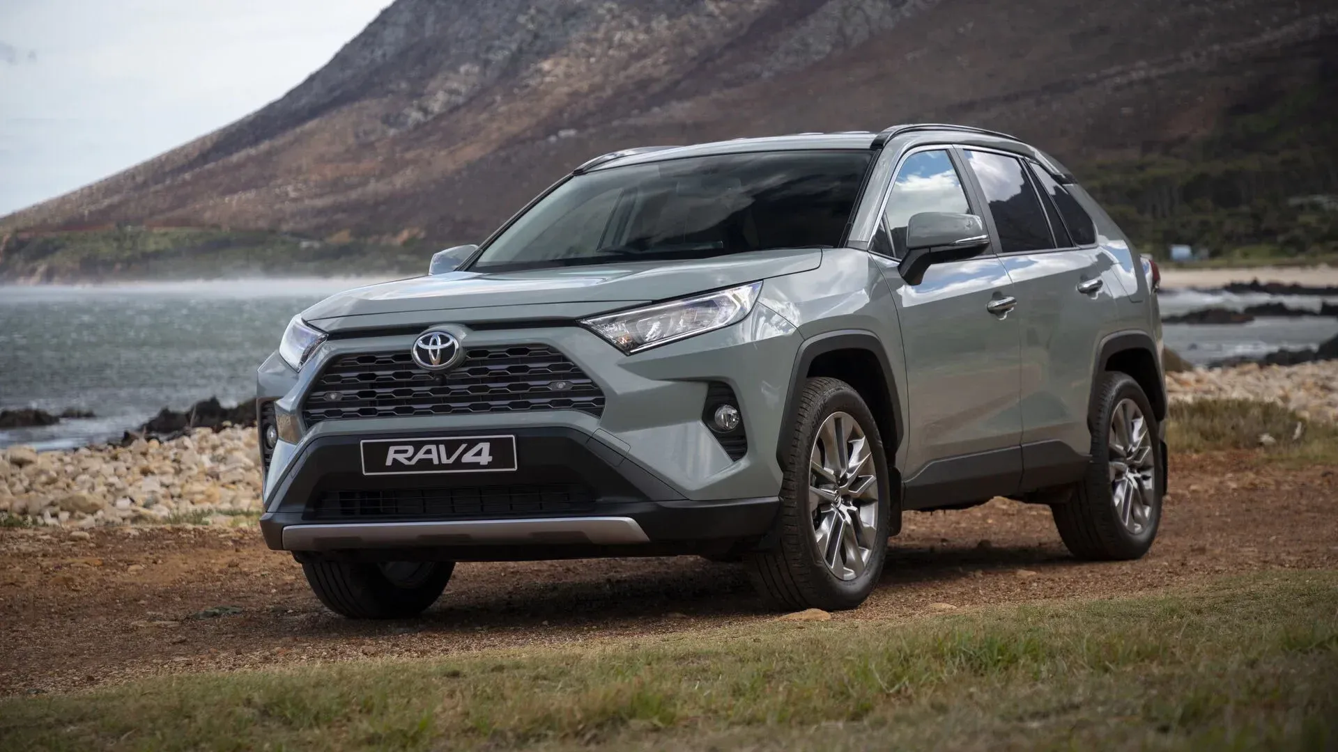 Gray Toyota RAV4 SUV parked on a rocky shore with mountains and water in the background