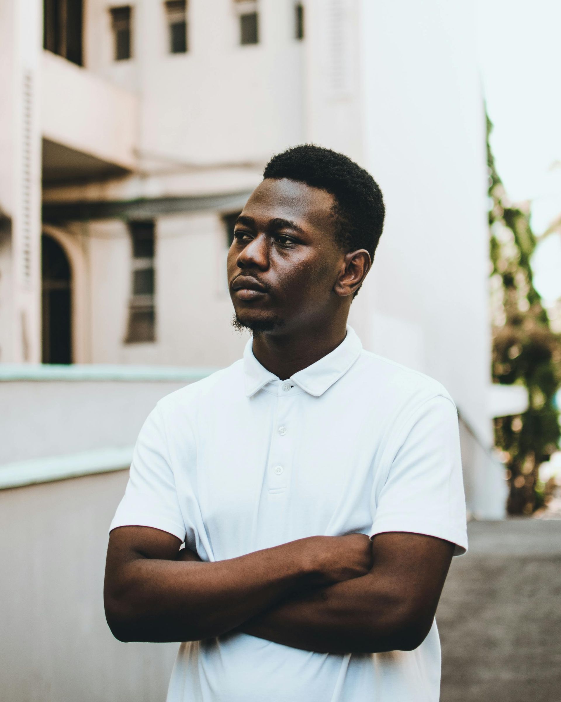 Man in a white polo shirt standing with arms crossed outdoors beside a building