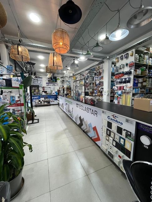 Pasillo luminoso de una tienda de electrónica con faroles colgantes, suelo blanco y estanterías repletas de aparatos y accesorios.