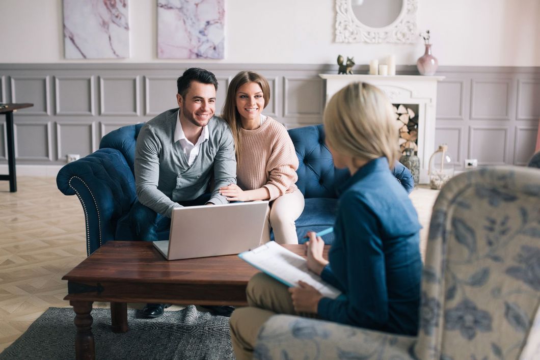 Two people on a couch meeting with a woman across a coffee table, using a laptop in a bright living room