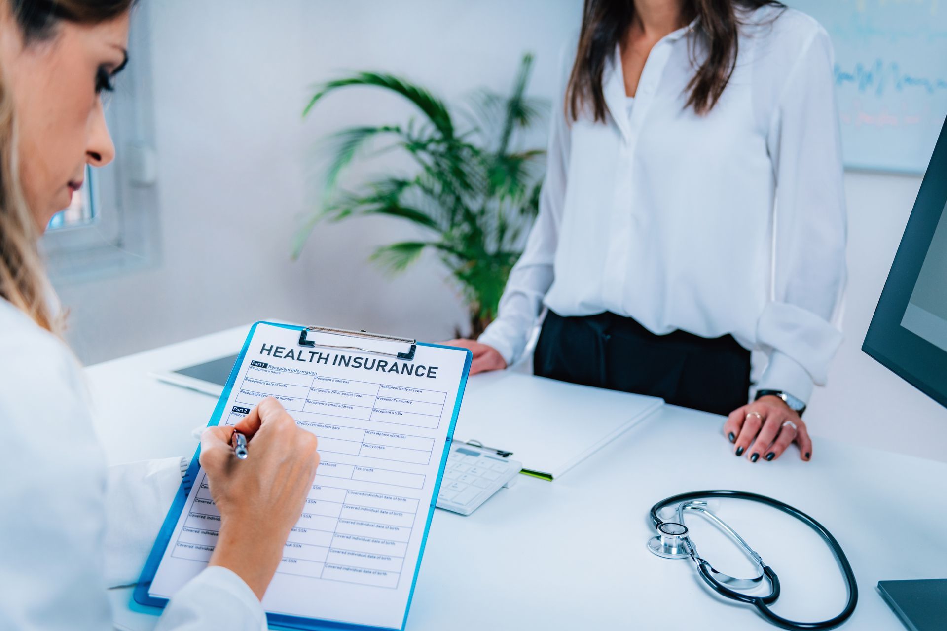 Doctor taking notes during a consultation with a patient at a white desk, stethoscope nearby.