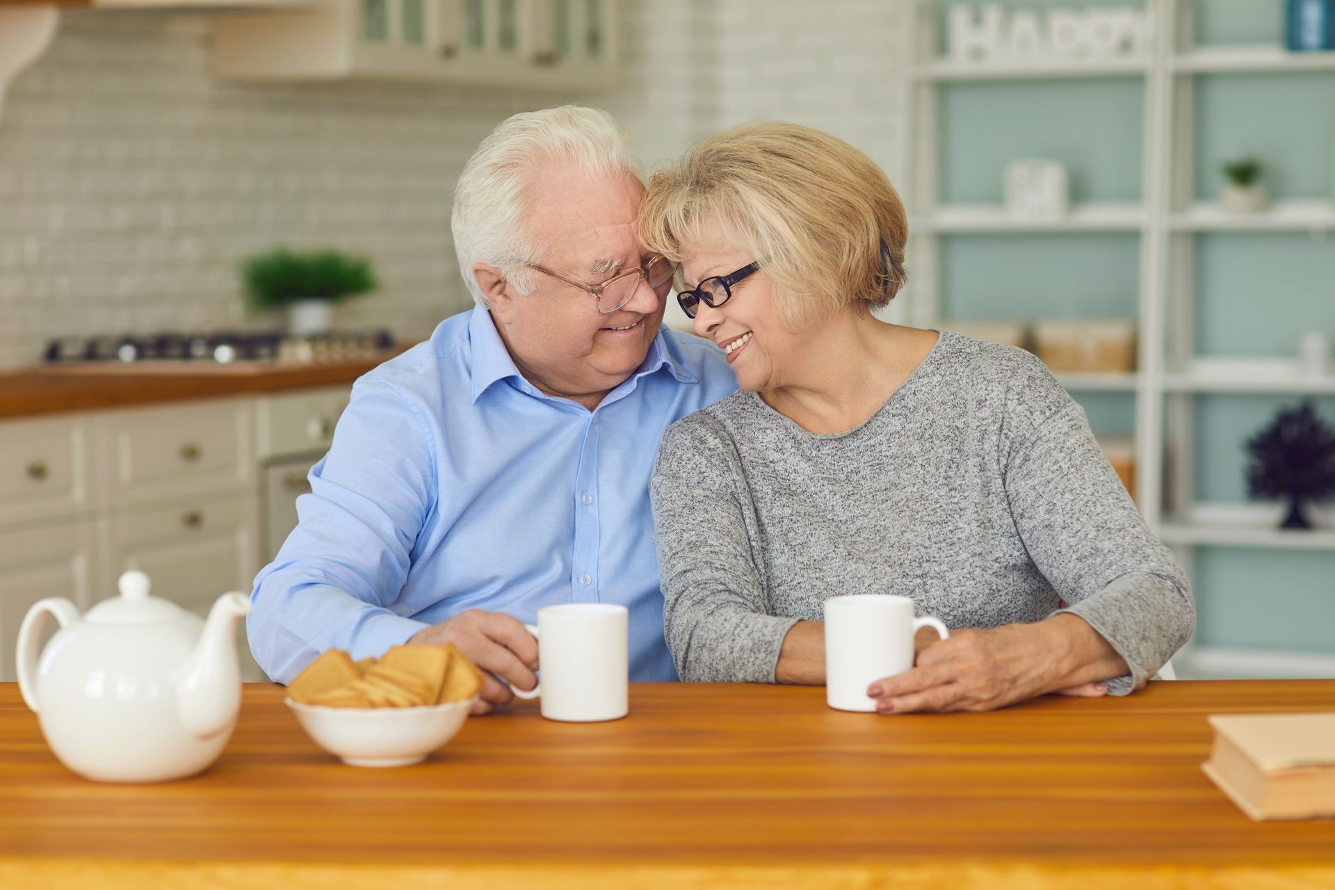 Two people sit close at a kitchen table, smiling and holding mugs beside tea and pastries.