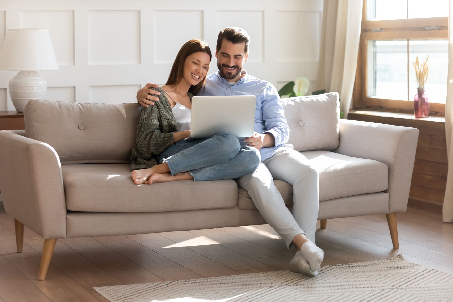 Couple sitting on a beige sofa using a laptop in a bright living room